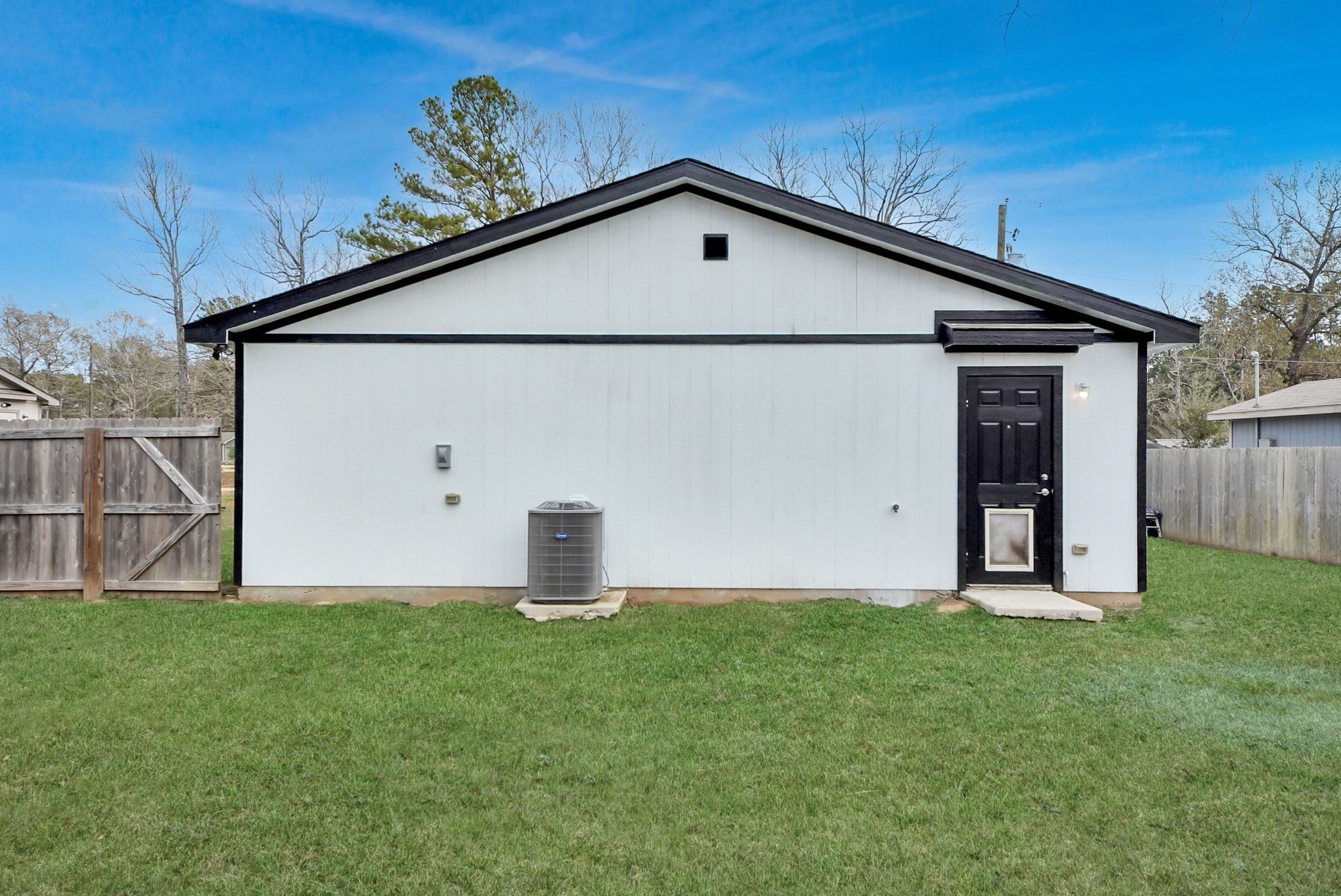 16124 Brettwood Lane Splendora, TX 77372 - Photo 33 of 40 This image shows the side of a house with a simple, clean white exterior and contrasting black trim. There’s a black door with a pet door and a central air conditioning unit. The yard is fenced and features green grass.