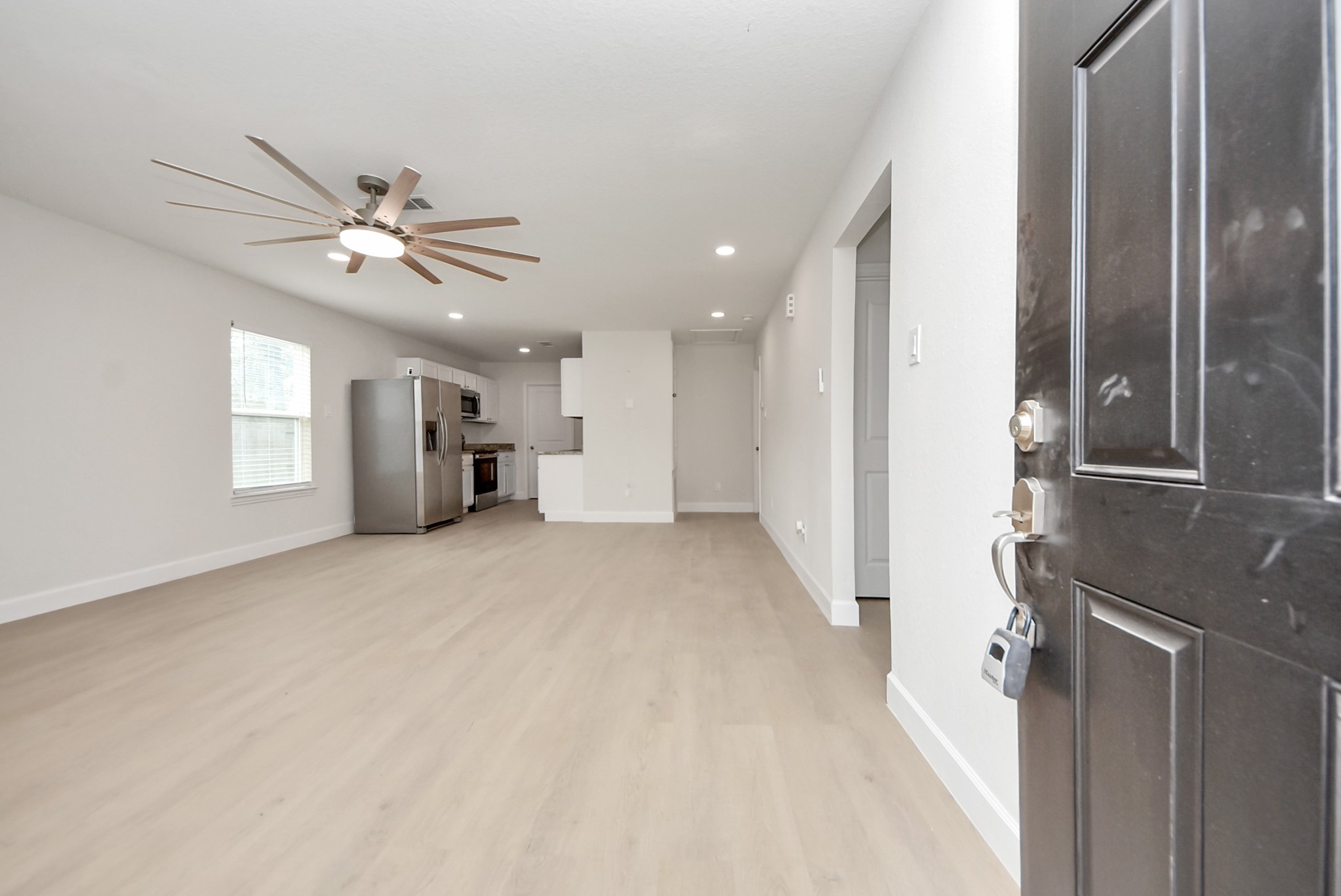 16124 Brettwood Lane Splendora, TX 77372 - Photo 4 of 40 This photo shows a modern, open-concept living area with light wood flooring, a ceiling fan, and large windows. The kitchen features stainless steel appliances and white cabinetry, creating a bright and inviting space.
