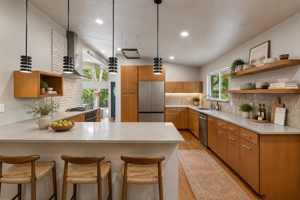 13321 Purple Sage Road Dallas, TX 75240 - Photo 6 of 30 AI Augmented Spacious kitchen featuring an expansive island with seating, stainless steel appliances, wood-finish cabinetry, light-toned countertops, and a brick-patterned backsplash