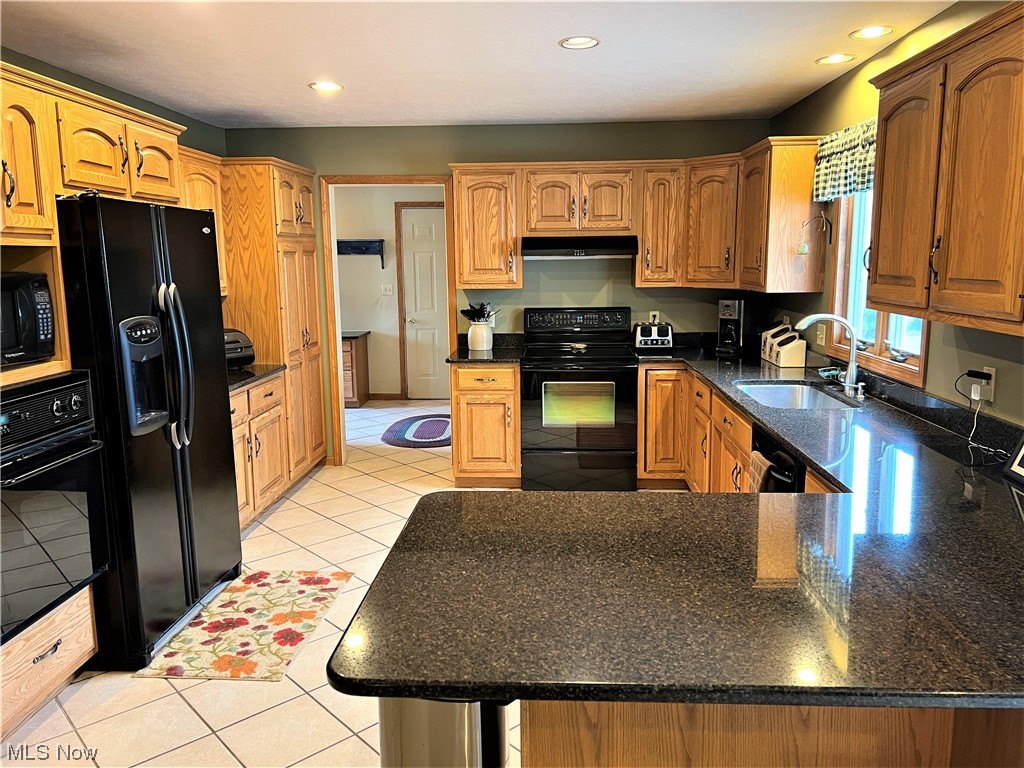 Kitchen with light tile floors, dark stone counters, black appliances, and brown cabinets
