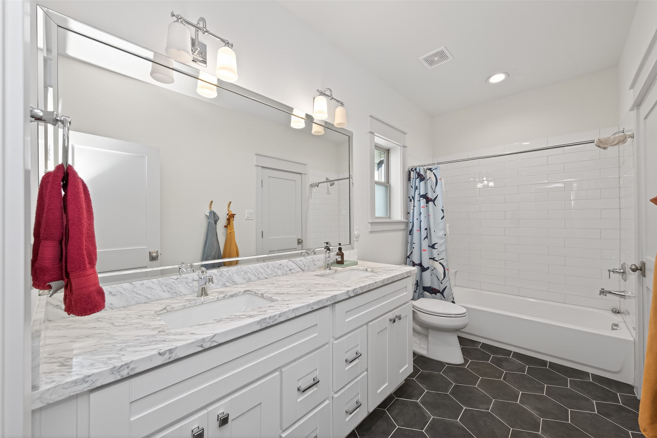 515 West 25th Street Houston, TX 77008 - Photo 21 of 35 This 6’2” x 12’5” secondary bathroom, shared between the two secondary bedrooms, features ceramic hex tile flooring, double sinks with a Carrara Marble countertop, a beveled mirror, Kohler toilet, and Park Harbor polished chrome two-bulb vanity lights. The tub/shower combo includes a 60” x 32” tub with subway tile surround and a polished chrome Signature Hardware tub and shower trim kit. Shaker-style cabinetry offers soft-close drawers and doors with polished chrome pull handles, and square knobs.