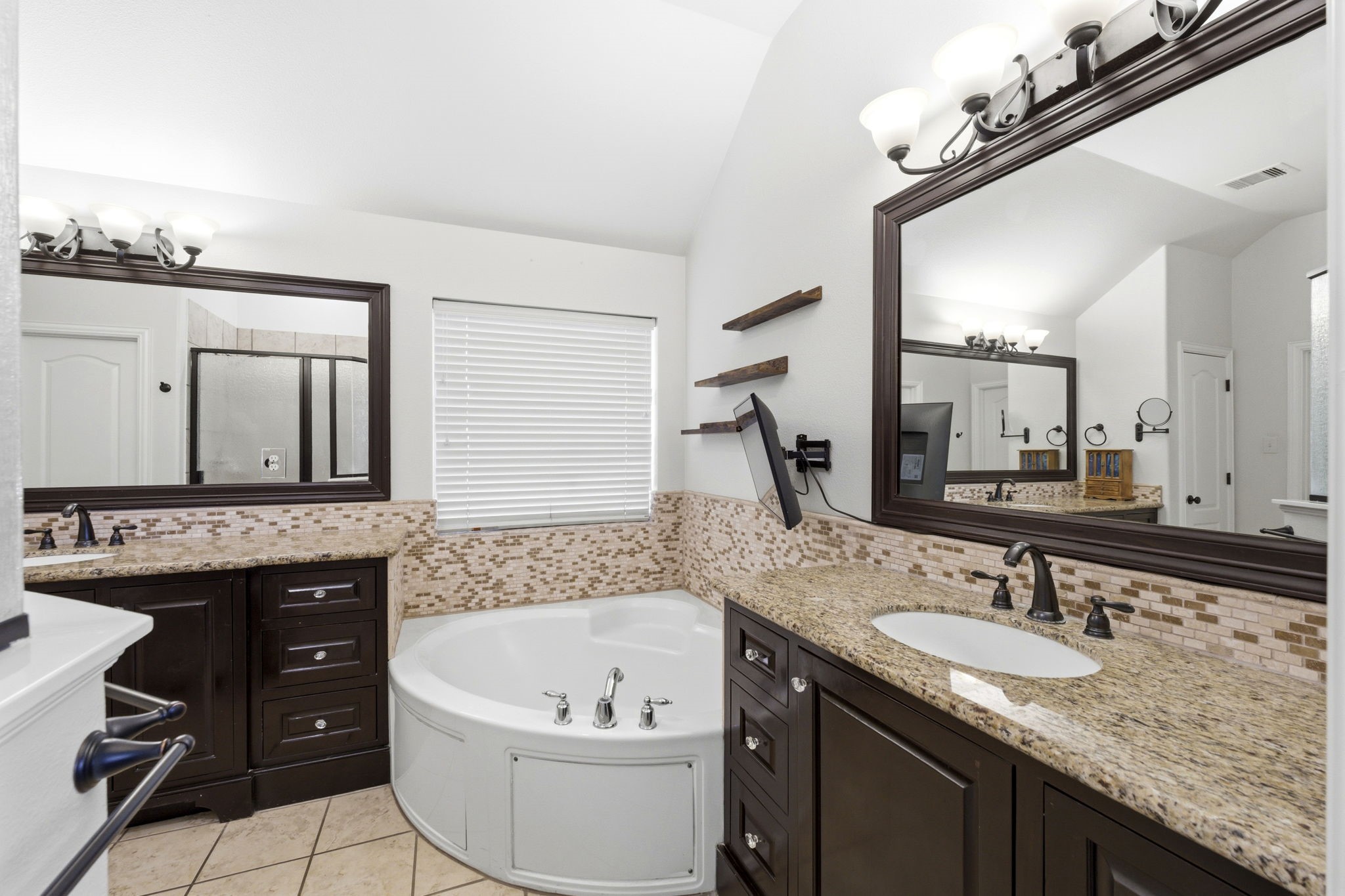 27502 Chris Ridge Court Spring, TX 77386 - Photo 18 of 37 This bathroom features a luxurious design with a large soaking tub, granite countertops, dual sinks, and elegant dark wood cabinetry. It includes a spacious mirror, stylish light fixtures, and a separate shower area, all complemented by a neutral color palette and tiled backsplash.