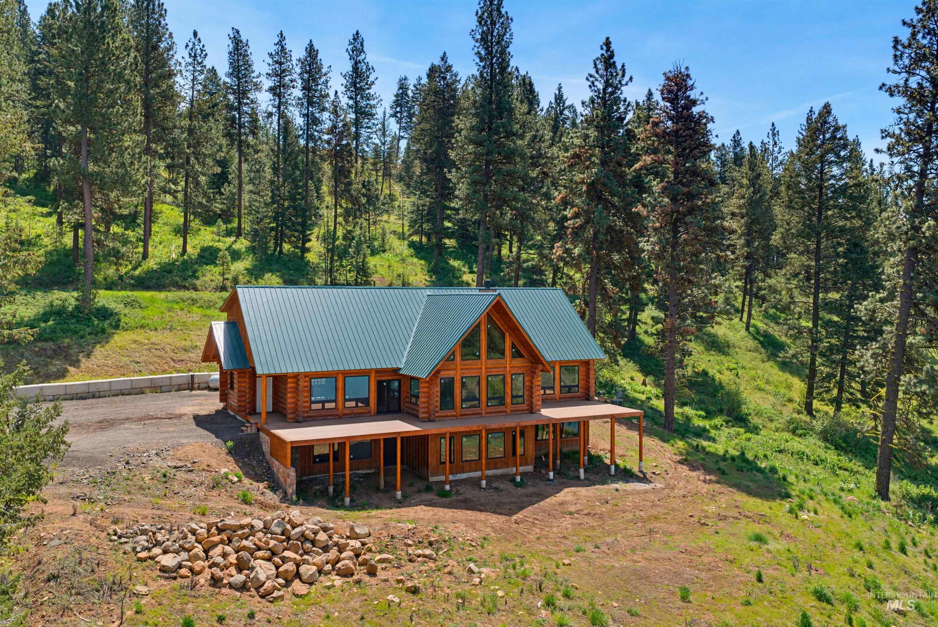 3322 Big Creek Road New Meadows, ID 83654 - Photo 9 of 50 Log cabin exterior featuring a green metal roof, a full-length covered porch, a prominent gable with large windows, and a surrounding natural landscape of evergreen trees and hillside vegetation