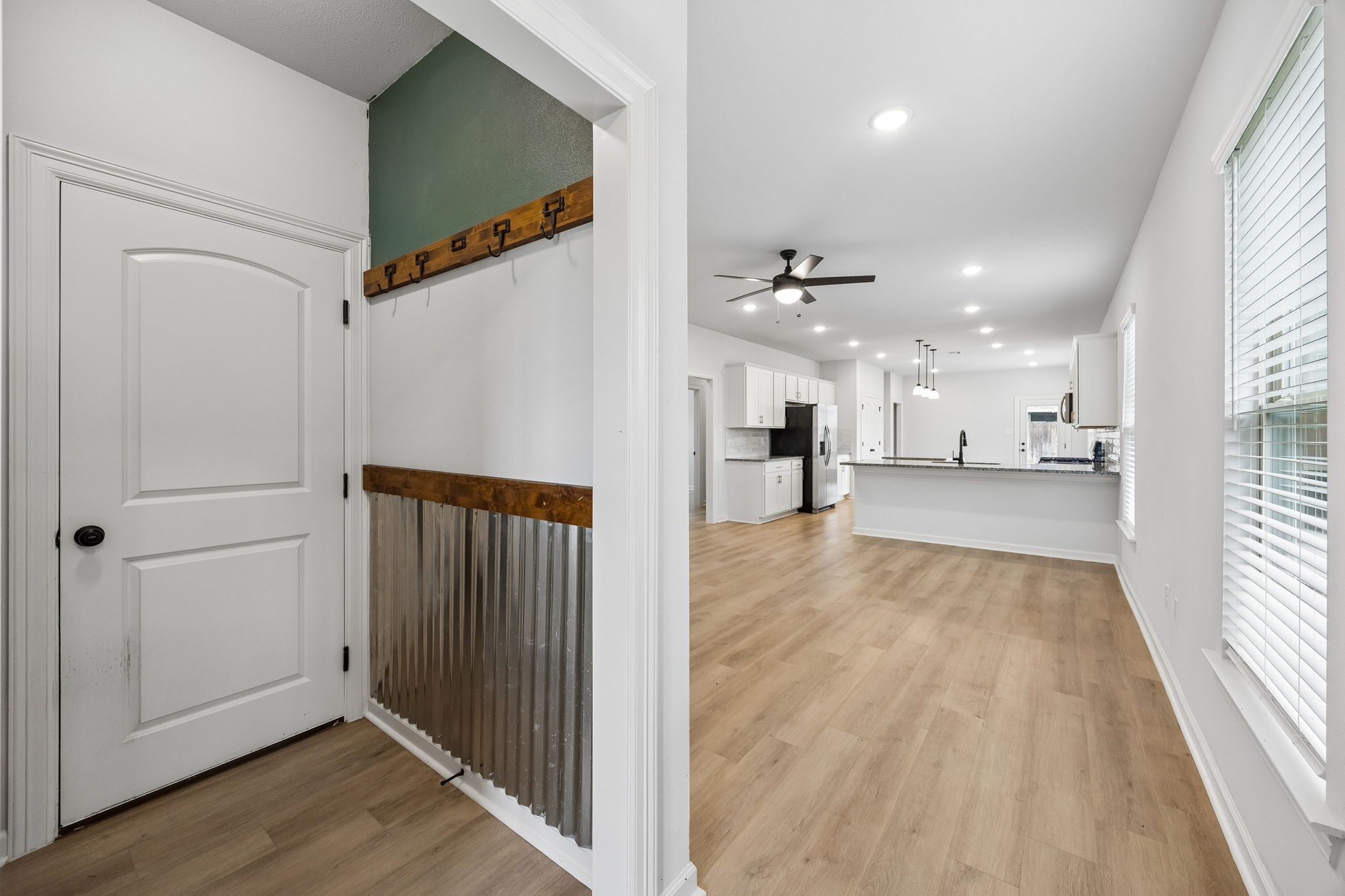 10902 Bonfire Trail Beasley, TX 77417 - Photo 26 of 36 This photo showcases a modern, open-concept living space with light vinyl plank floors and a bright, airy feel. The kitchen features white cabinetry, stainless steel appliances, and a large island. A mudroom area with rustic hooks and corrugated metal wainscoting is visible near the entryway.