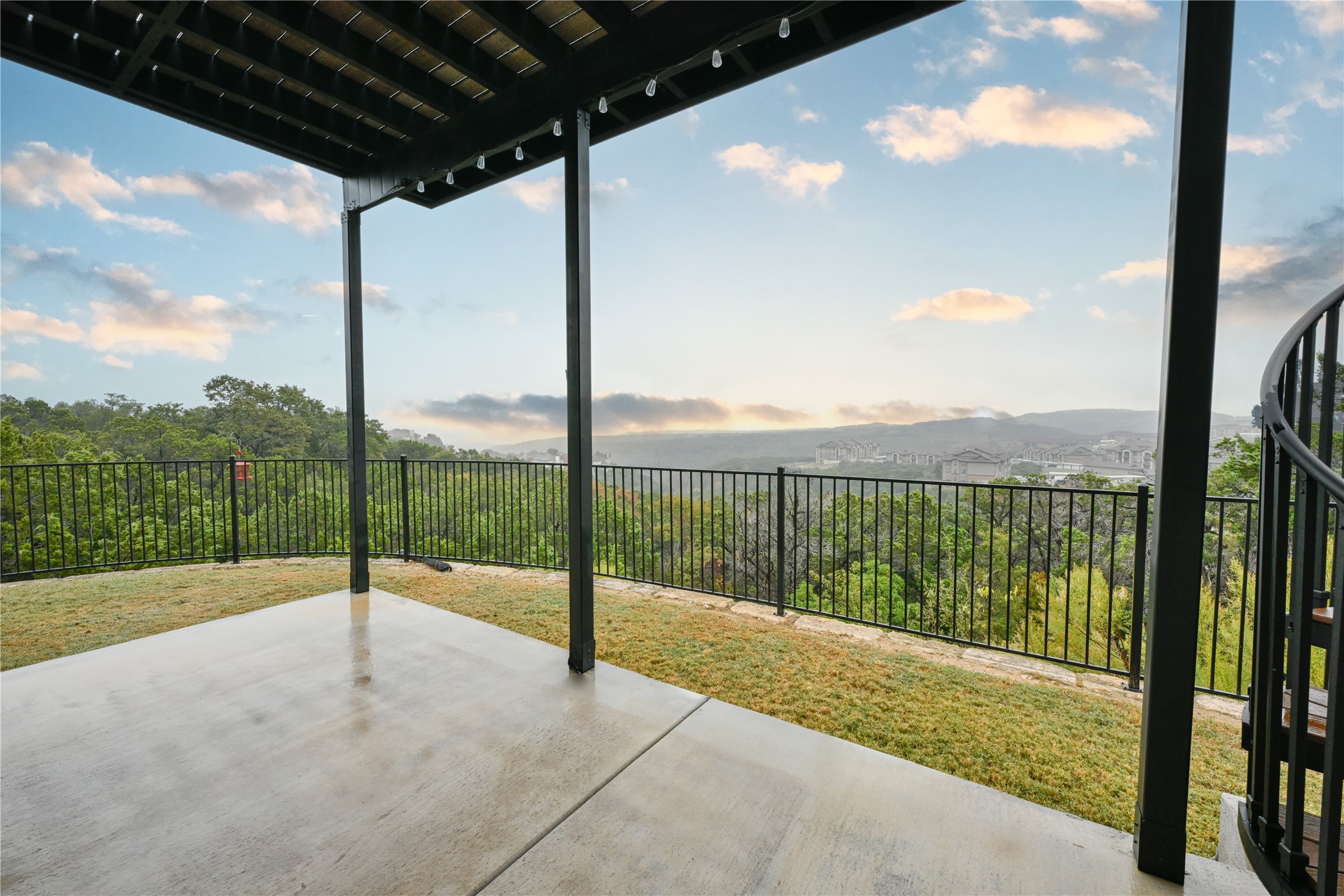 232 Sunrise Ridge Loop Austin, TX 78738 - Photo 35 of 37 Covered concrete patio with a spiral staircase, black metal railing, overhead string lighting, and panoramic distant hill and cityscape