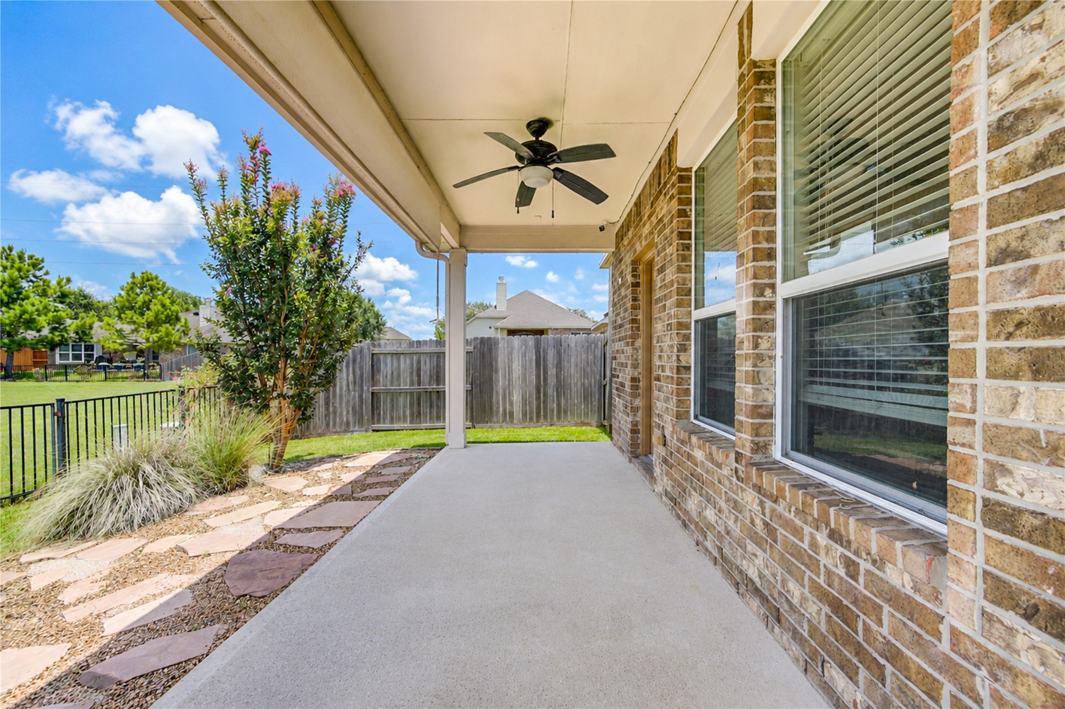 6014 Watford Bend Rosenberg, TX 77471 - Photo 22 of 24 Spacious covered back patio featuring a smooth concrete pad, ceiling fan, and classic brick exterior walls. The outdoor living area overlooks a fully fenced backyard with a landscaped flagstone stepping-stone pathway, decorative rock borders, flowering crape myrtle, and lush green grass. Perfect space for outdoor dining, relaxing, and entertaining guests.