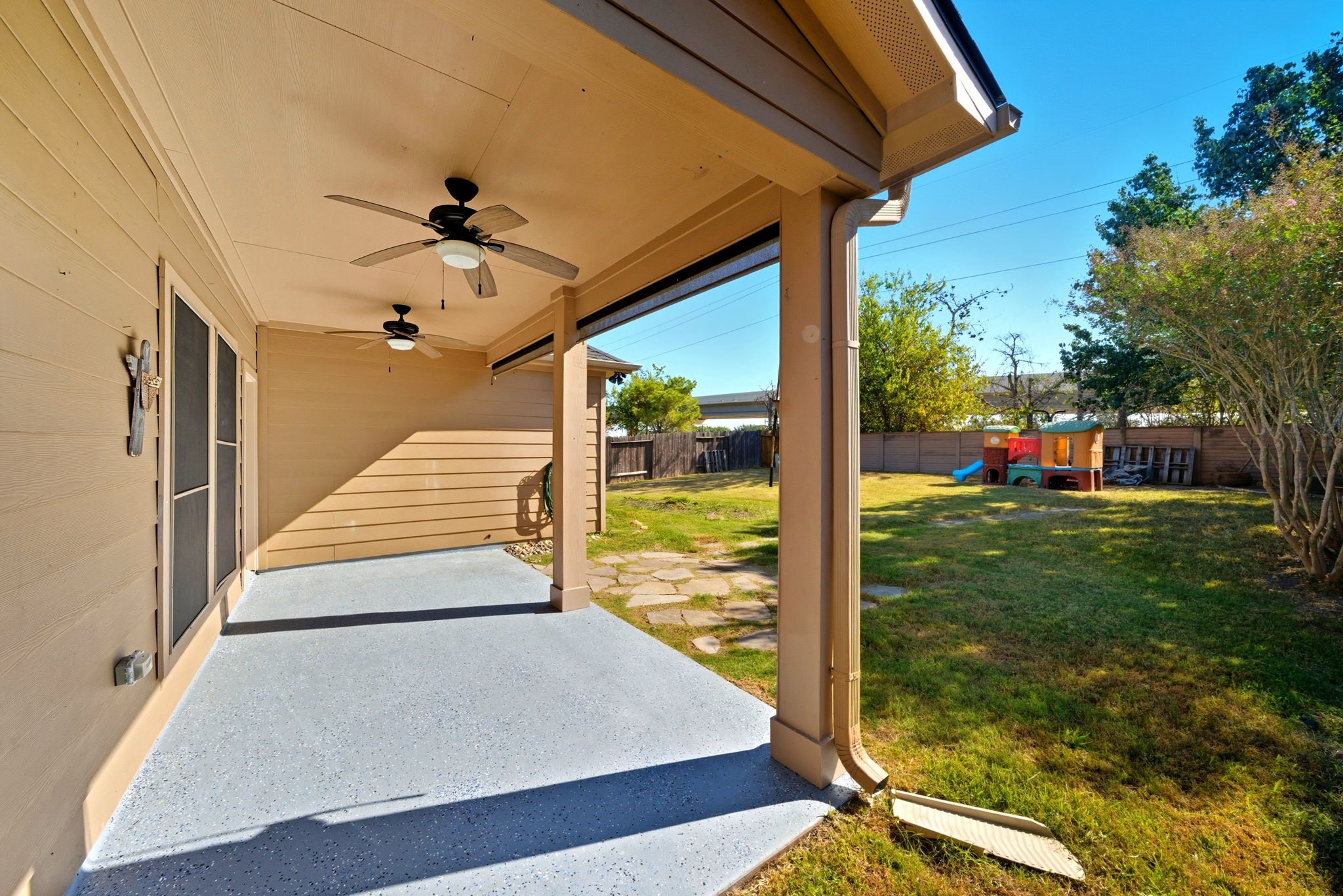 23139 Tranquil Spring Lane Katy, TX 77494 - Photo 21 of 22 Home showcases a spacious backyard with a covered patio, featuring ceiling fans for comfort. The area overlooks a grassy yard with mature trees and a children's play structure, ideal for outdoor activities and relaxation.