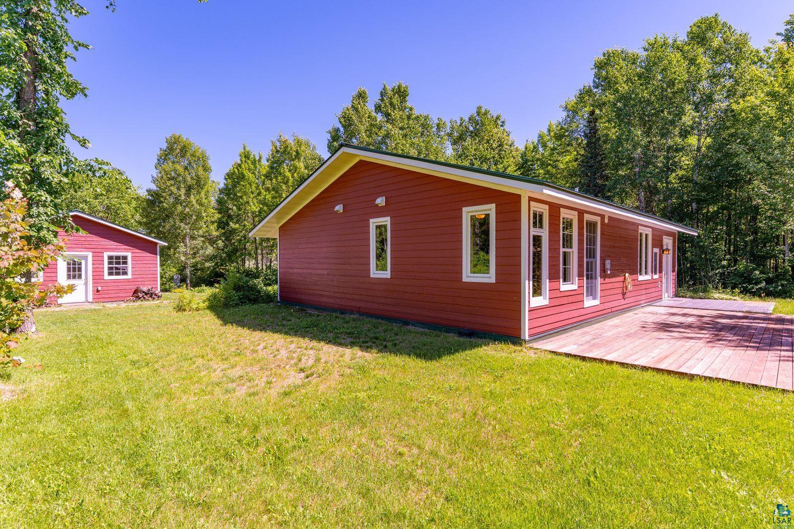 9734 Alango Road Angora, MN 55703 - Photo 23 of 41 Red siding structure featuring multiple windows, a long wooden deck, and a separate red outbuilding with a white door and window, all surrounded by mature trees