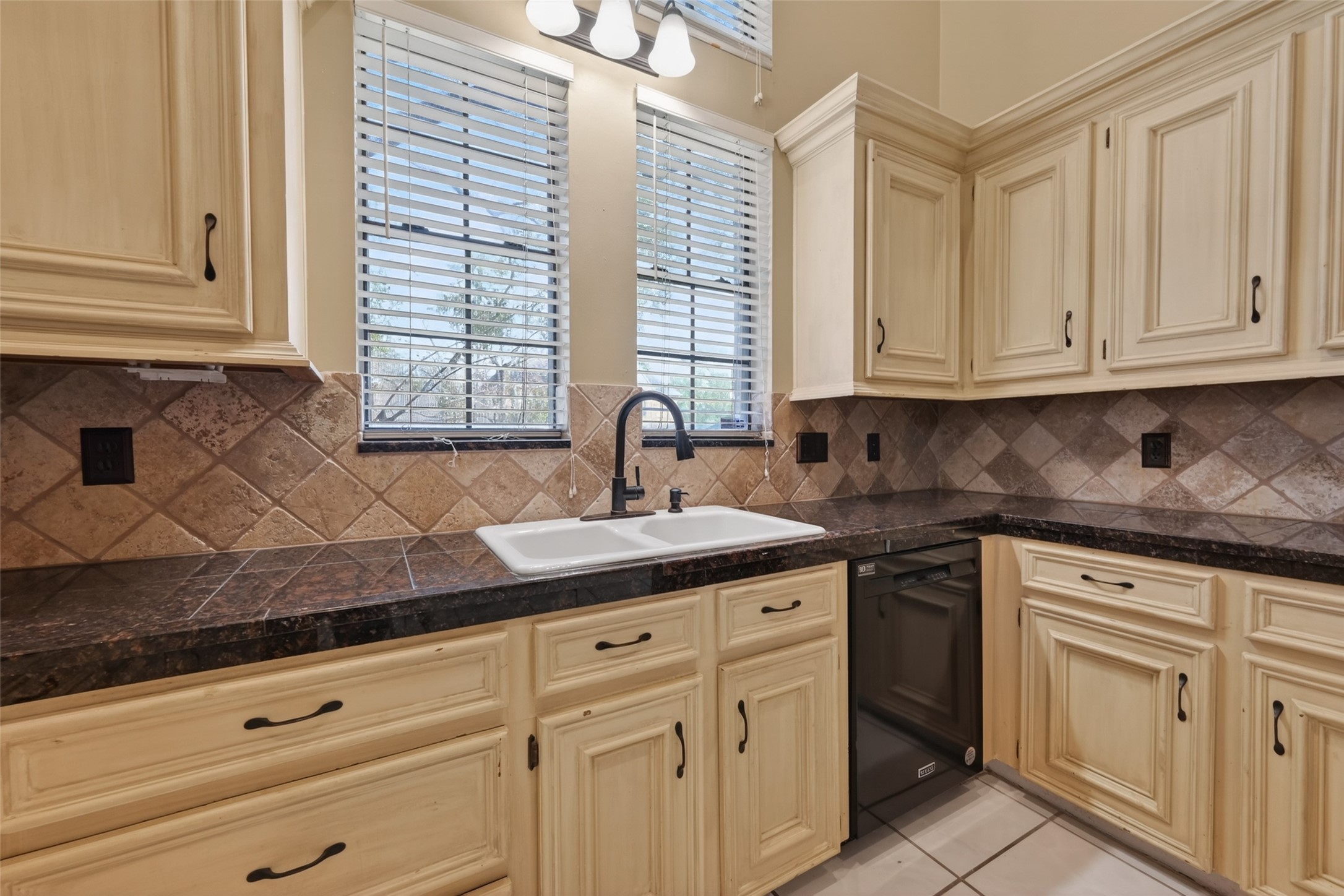 5802 Ashmere Lane Spring, TX 77379 - Photo 11 of 41 This kitchen features cream-colored cabinetry with classic hardware, dark granite countertops, and a double sink under double windows. The backsplash is tiled in a warm, earthy tone, and the space includes a built-in dishwasher.