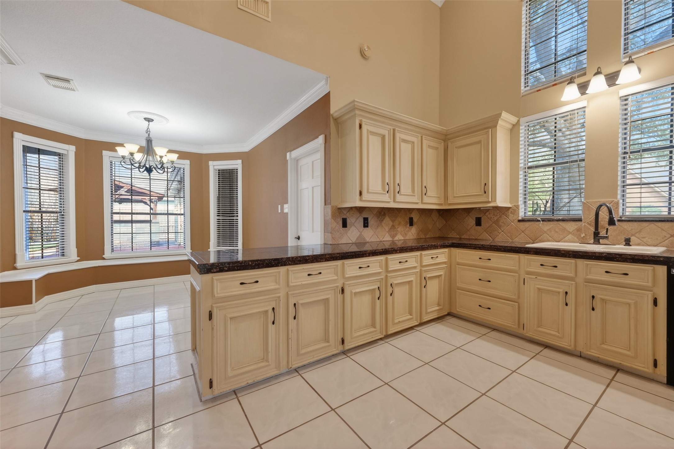5802 Ashmere Lane Spring, TX 77379 - Photo 12 of 41 This spacious kitchen features cream cabinetry with dark countertops, a tiled backsplash, and large windows for natural light. The adjacent dining area has a bay window with a built-in bench and a stylish chandelier, all set on a glossy tile floor.