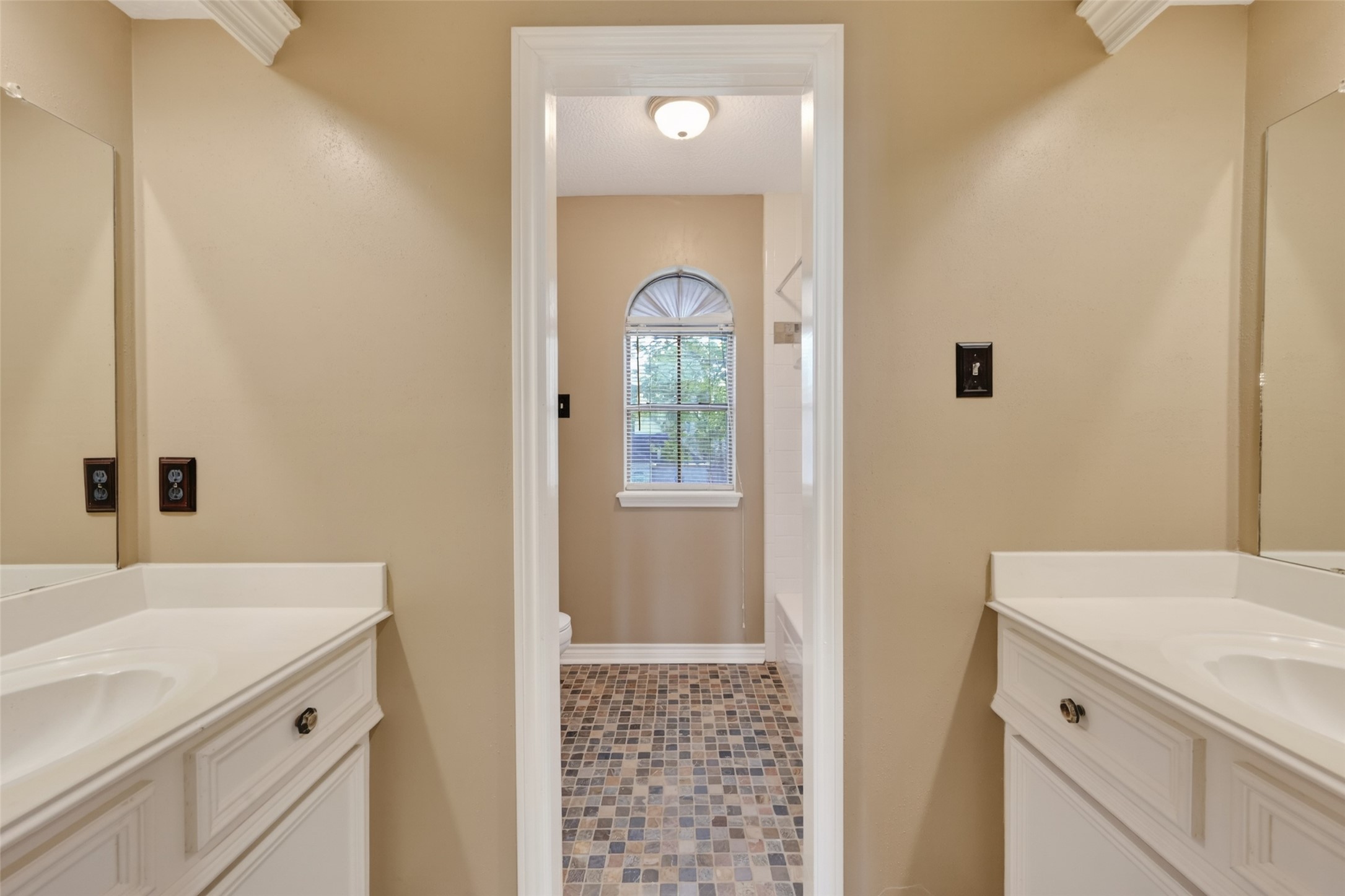 5802 Ashmere Lane Spring, TX 77379 - Photo 29 of 41 This photo shows a bright bathroom with dual vanities featuring white countertops. The room has beige walls and a tile floor. A window with a decorative arch lets in natural light, and a doorway leads to a shower area.