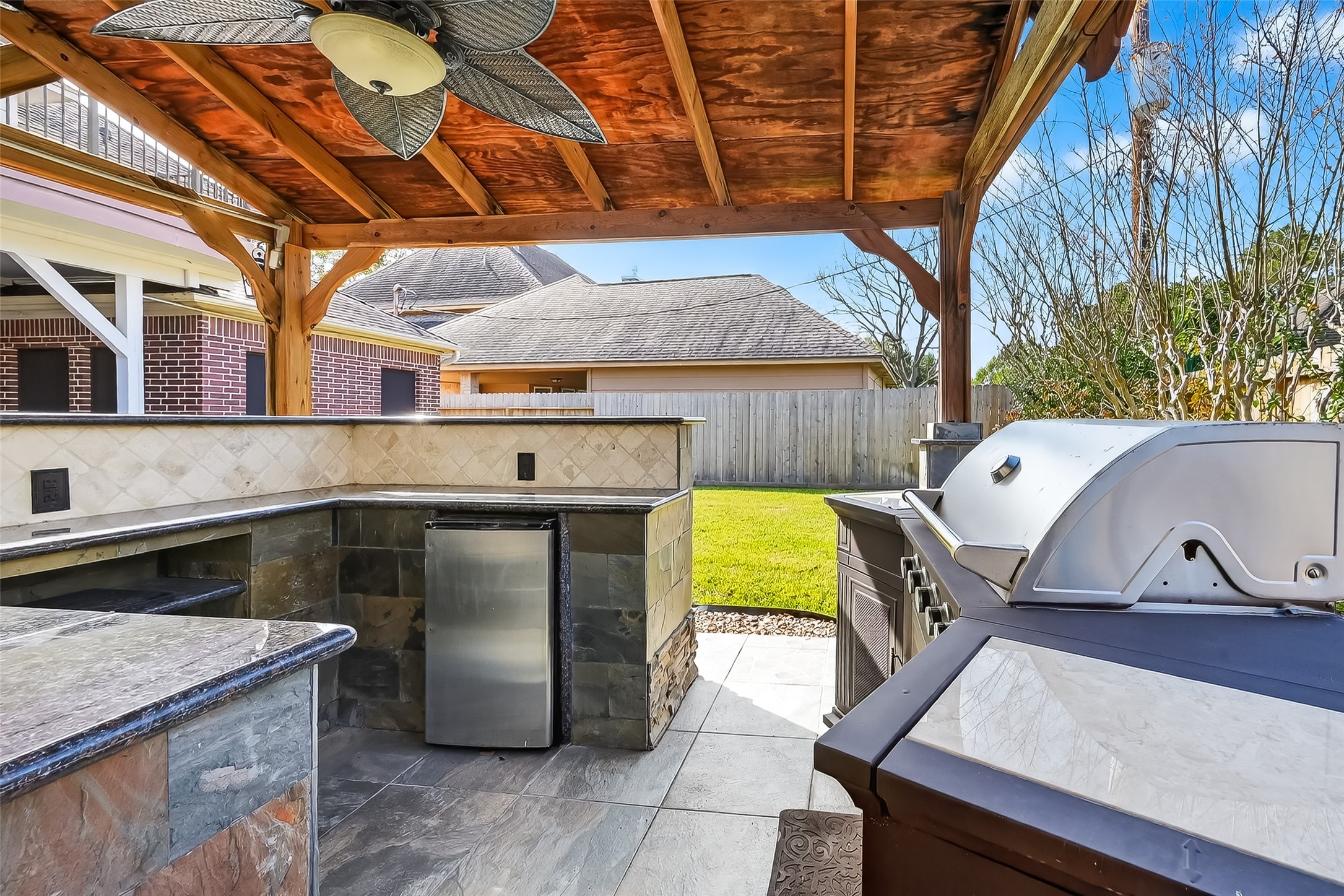 5802 Ashmere Lane Spring, TX 77379 - Photo 38 of 41 This photo showcases a stylish outdoor kitchen with a granite countertop, built-in grill, mini-fridge, and a ceiling fan under a wooden pergola, perfect for entertaining in a spacious backyard.