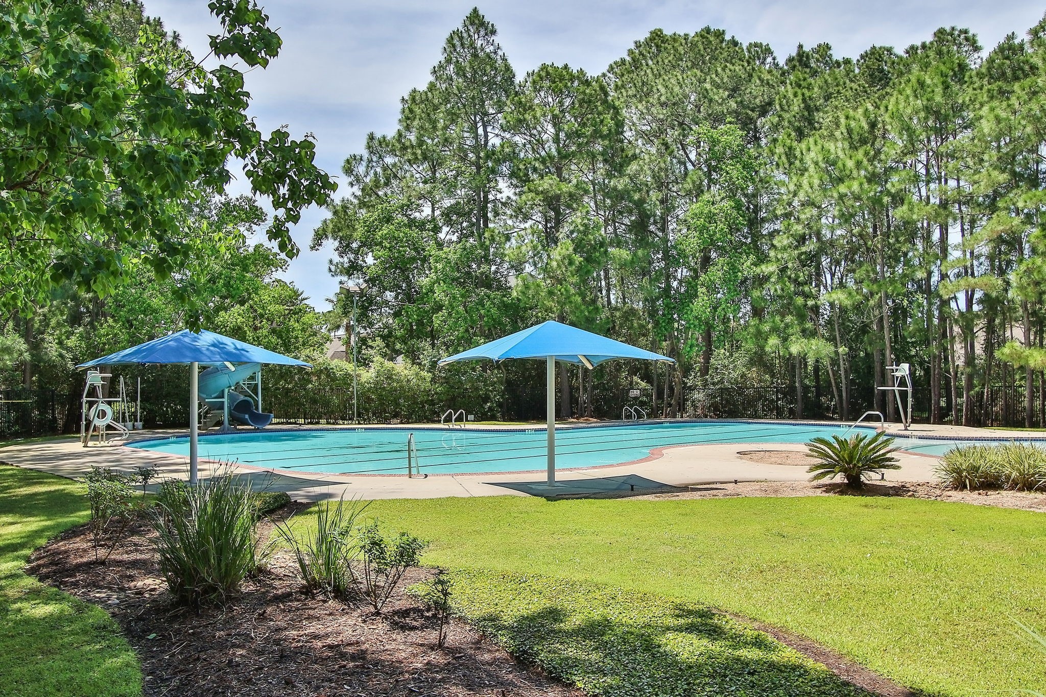 20410 Misty Crossing Lane Spring, TX 77379 - Photo 40 of 50 This photo showcases a serene outdoor pool area surrounded by lush greenery. The pool features a slide and is accompanied by blue sun umbrellas, offering a relaxing and inviting atmosphere.