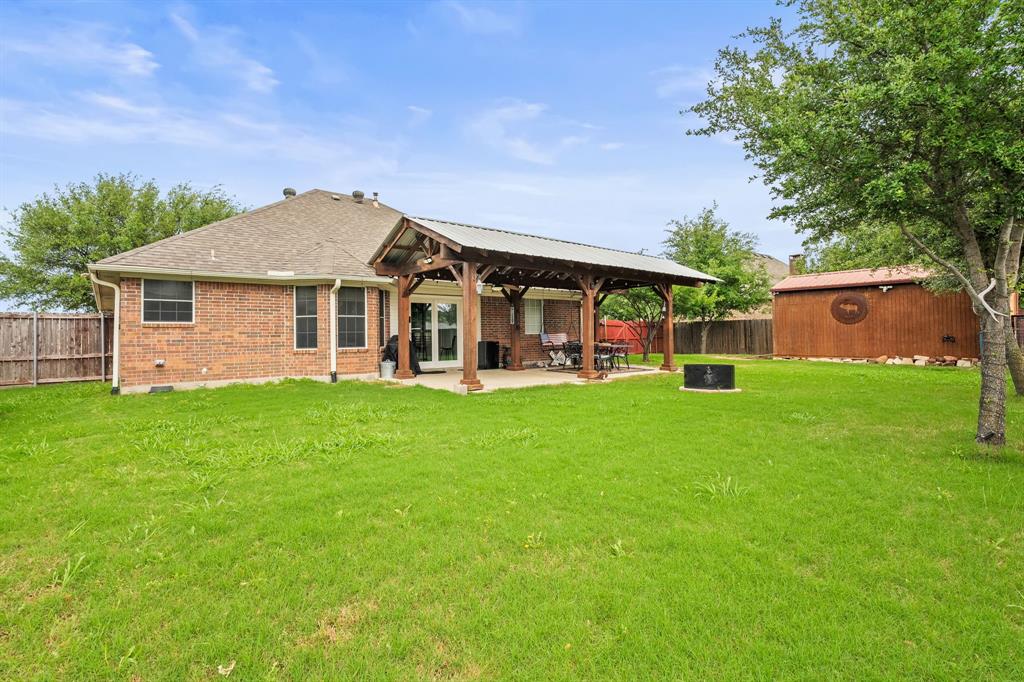 1401 Hickory Drive Pilot Point, TX 76258 - Photo 24 of 34 Expansive backyard featuring a covered patio with a corrugated metal roof, a wood-framed pergola structure, a large grassy area, mature trees, and a separate storage building with a metal roof