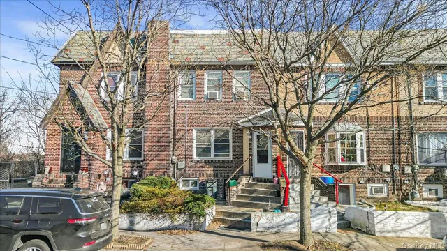 a front view of a building with large trees and cars parked on road