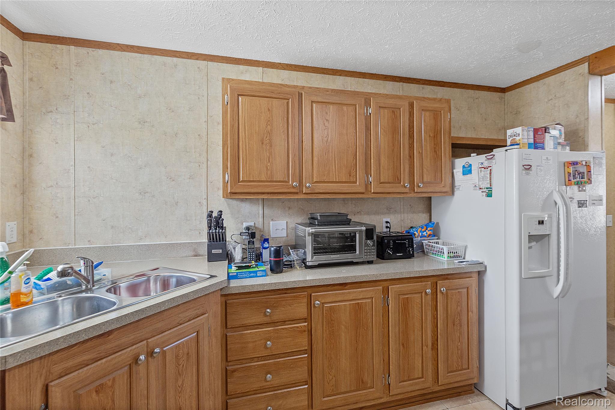 Kitchen with white refrigerator with ice dispenser, light countertops, brown cabinets, a textured ceiling, and crown molding