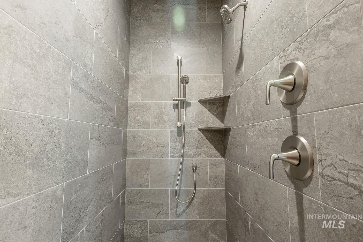 1171 West Recess Way Kuna, ID 83634 - Photo 13 of 28 Shower stall featuring large format grey tile, a rain shower head, a handheld shower fixture, and two built-in corner shelves