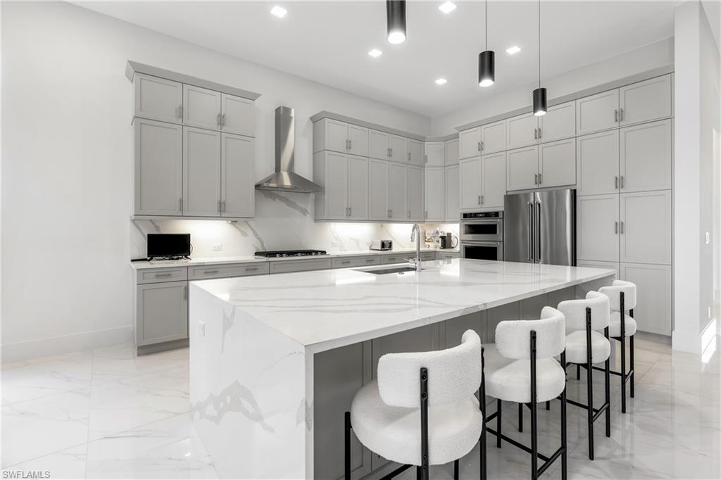 9675 Moonflower Court Naples, FL 34114 - Photo 7 of 32 Kitchen featuring gray cabinetry, light stone counters, wall chimney exhaust hood, a kitchen bar, and stainless steel appliances