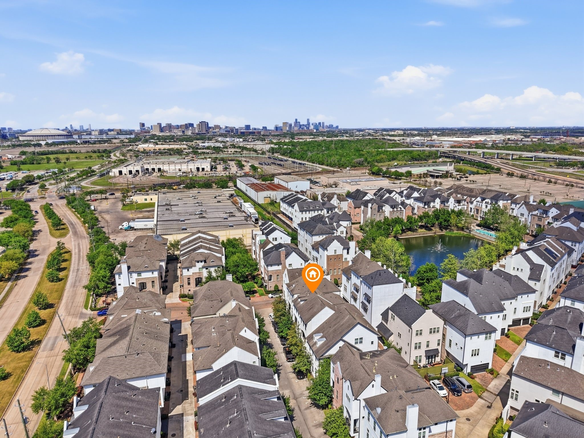 9427 London Bridge Station Houston, TX 77045 - Photo 28 of 30 Aerial view of a modern residential neighborhood featuring neatly arranged townhouses. The area is lush with greenery and has a pond, creating a serene environment. The city skyline is visible in the distance, indicating proximity to urban amenities.