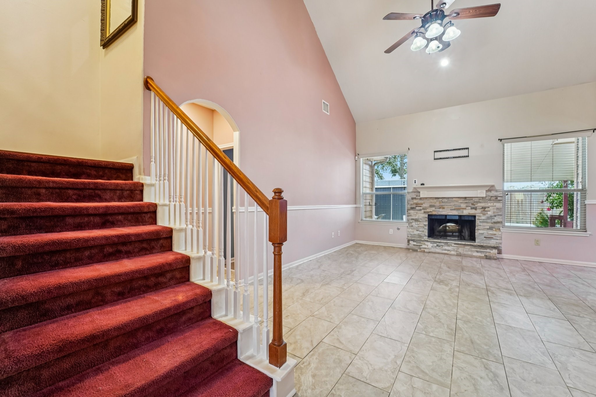 3818 Ridge Canyon Road Baytown, TX 77521 - Photo 14 of 42 This spacious living area features a vaulted ceiling with a ceiling fan, a cozy stone fireplace, and abundant natural light from two large windows. The room is complemented by tile flooring and an adjacent staircase with red carpet and white railings, adding a touch of elegance.