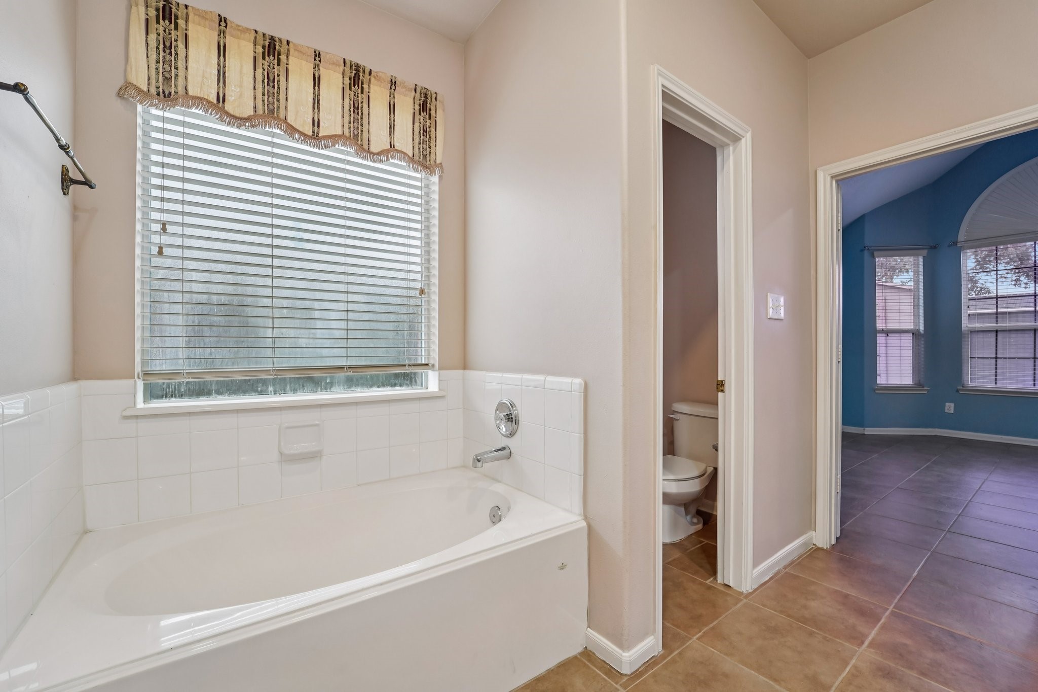3818 Ridge Canyon Road Baytown, TX 77521 - Photo 29 of 42 This photo features a bright bathroom with a large soaking tub and a window with blinds. Adjacent is a toilet area, and the space opens into a room with tiled flooring and blue walls, offering natural light through multiple windows.