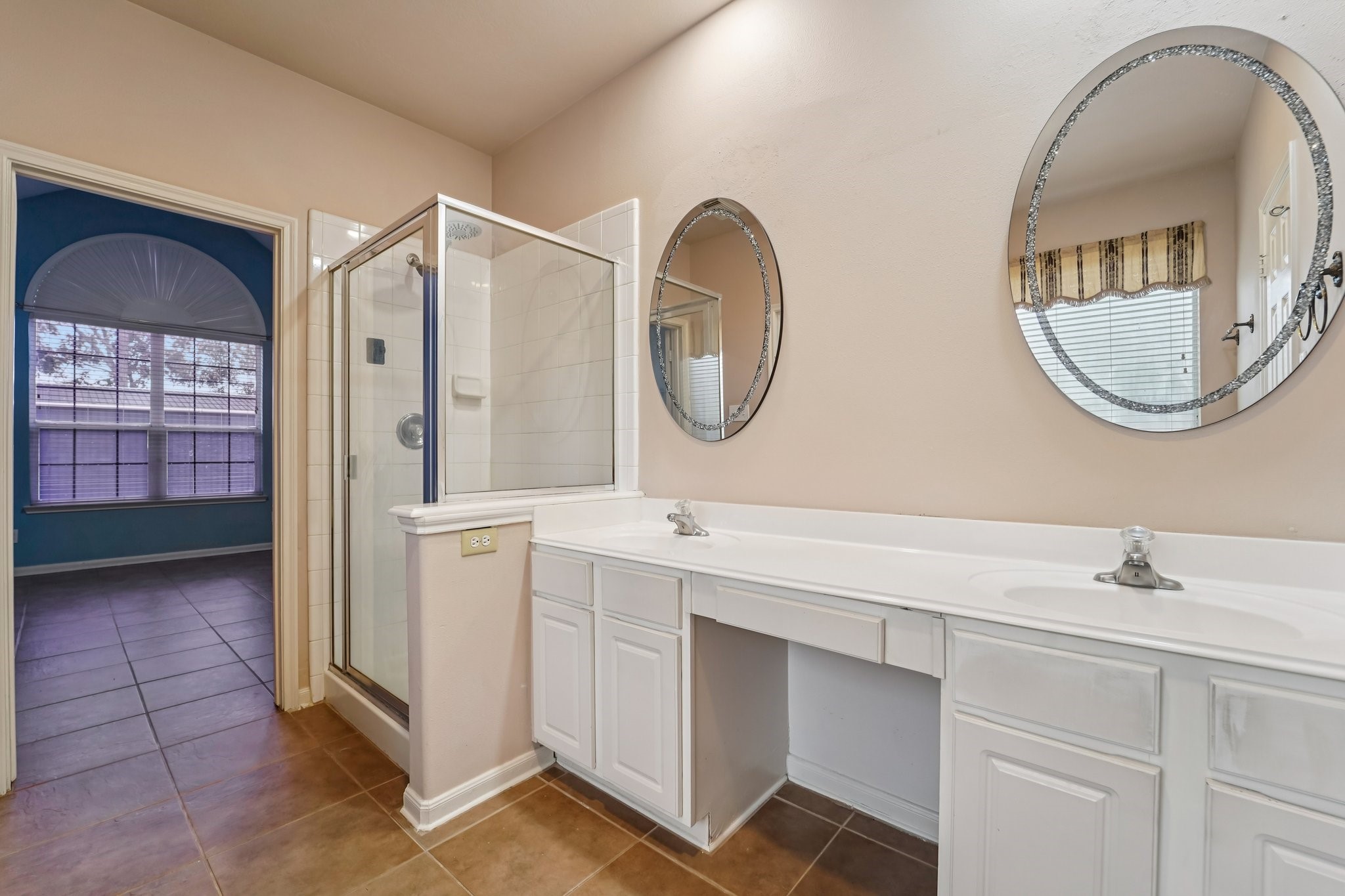 3818 Ridge Canyon Road Baytown, TX 77521 - Photo 30 of 42 The photo features a bright bathroom with dual sinks and round mirrors. It has a glass-enclosed shower and leads into a room with large windows, providing ample natural light. The neutral tones create a clean, modern feel.