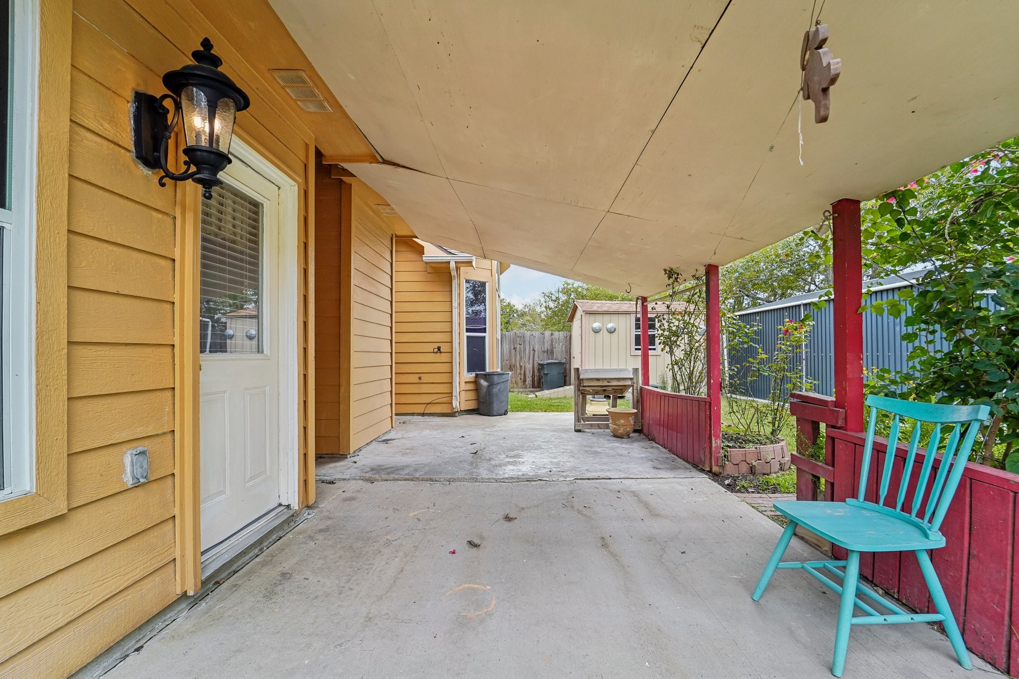3818 Ridge Canyon Road Baytown, TX 77521 - Photo 39 of 42 This photo shows a cozy covered patio area with a wooden exterior in a warm yellow hue. The space features a decorative outdoor lamp, a teal chair, and a small garden area with plants, offering a welcoming outdoor retreat.
