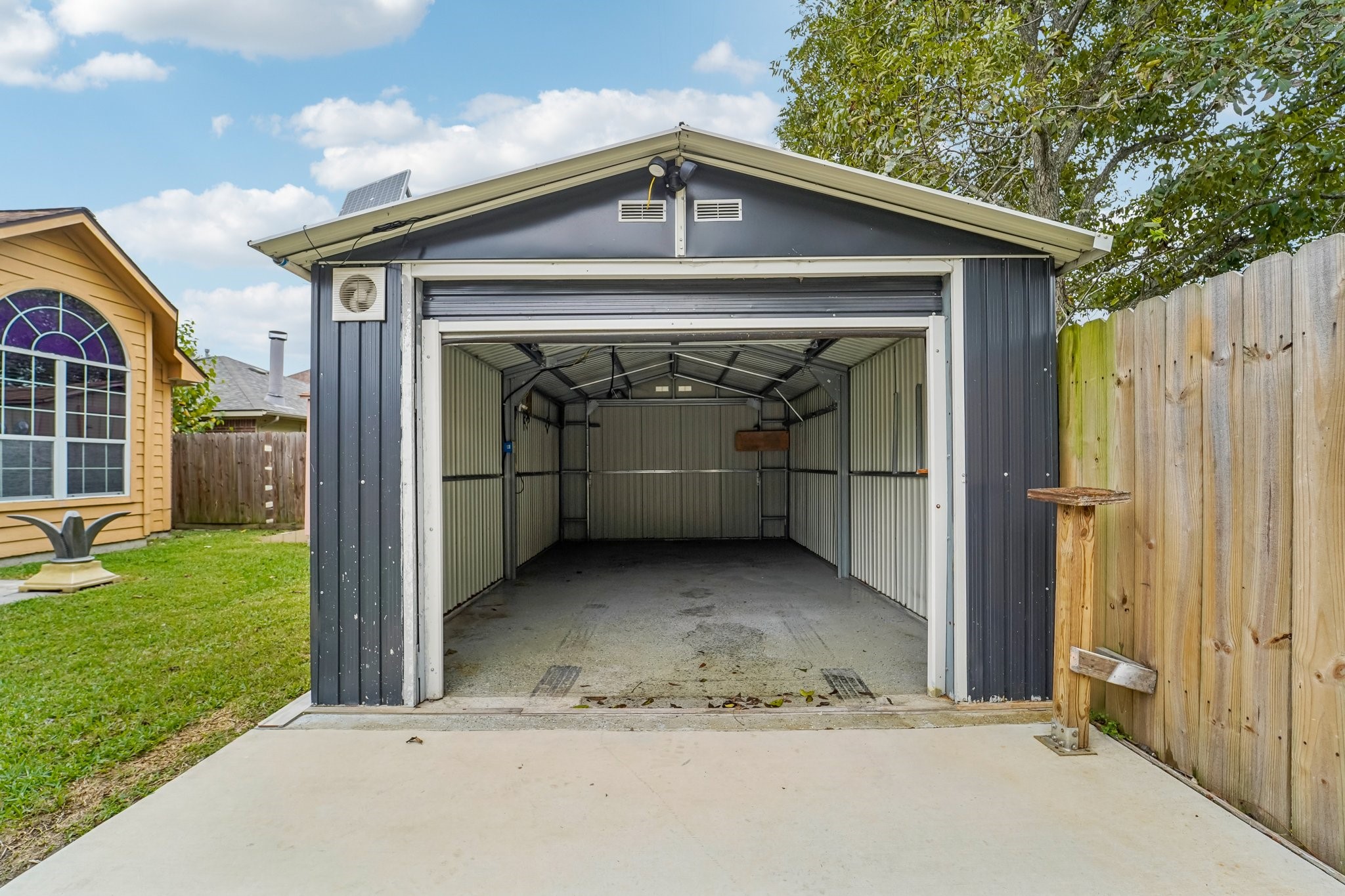 3818 Ridge Canyon Road Baytown, TX 77521 - Photo 4 of 42 This photo shows a detached garage with a metal exterior, situated in a backyard. It has a roll-up door and is adjacent to a wooden fence. The garage is surrounded by a grassy area and other neighboring structures.