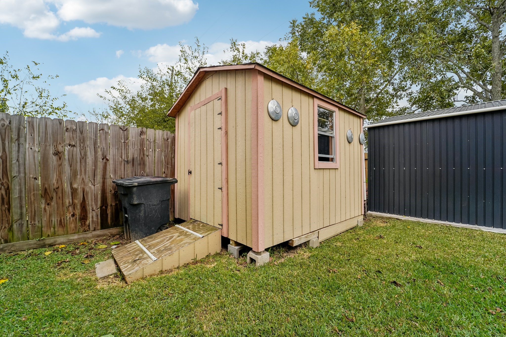 3818 Ridge Canyon Road Baytown, TX 77521 - Photo 42 of 42 This photo shows a small, wooden storage shed in a backyard. The shed is next to a tall wooden fence and a larger metal structure. It features a sloped roof, a small window, and a ramp leading to the door, set on a grassy lawn.