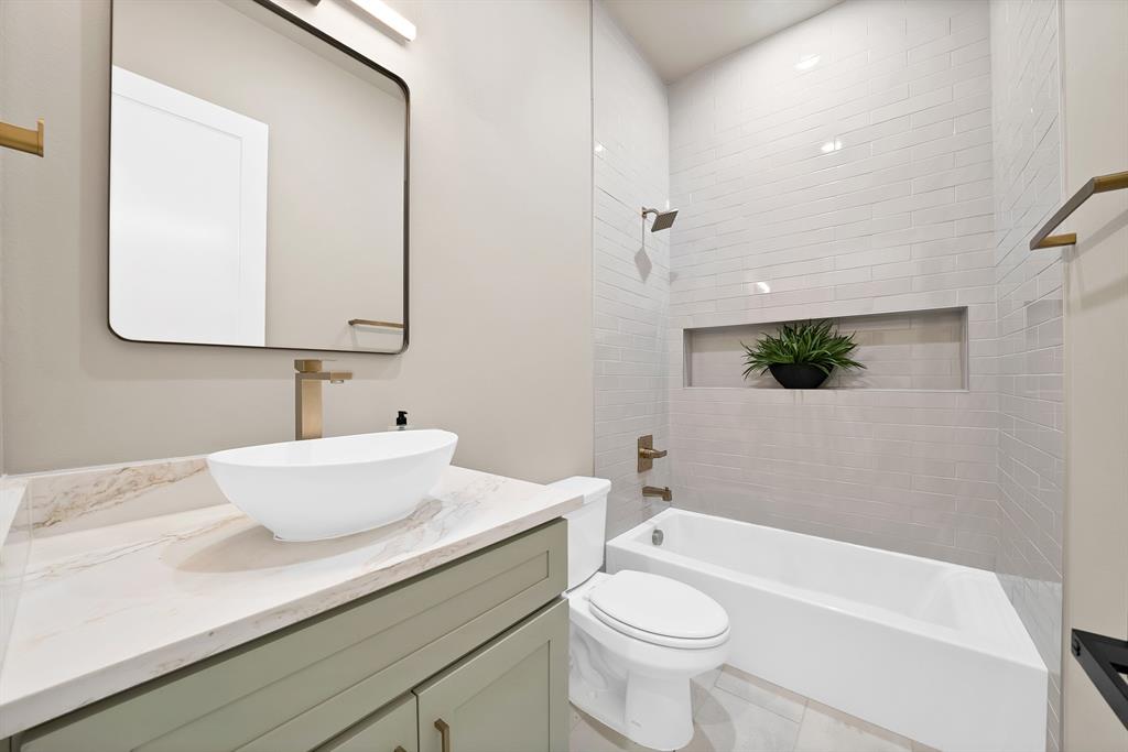 1415 Fayette Street Dallas, TX 75203 - Photo 20 of 40 White vessel sink with brass-finish faucet, framed mirror with integrated lighting, green-finish vanity with brass-finish hardware, subway-tiled shower with recessed niche, and a white bathtub