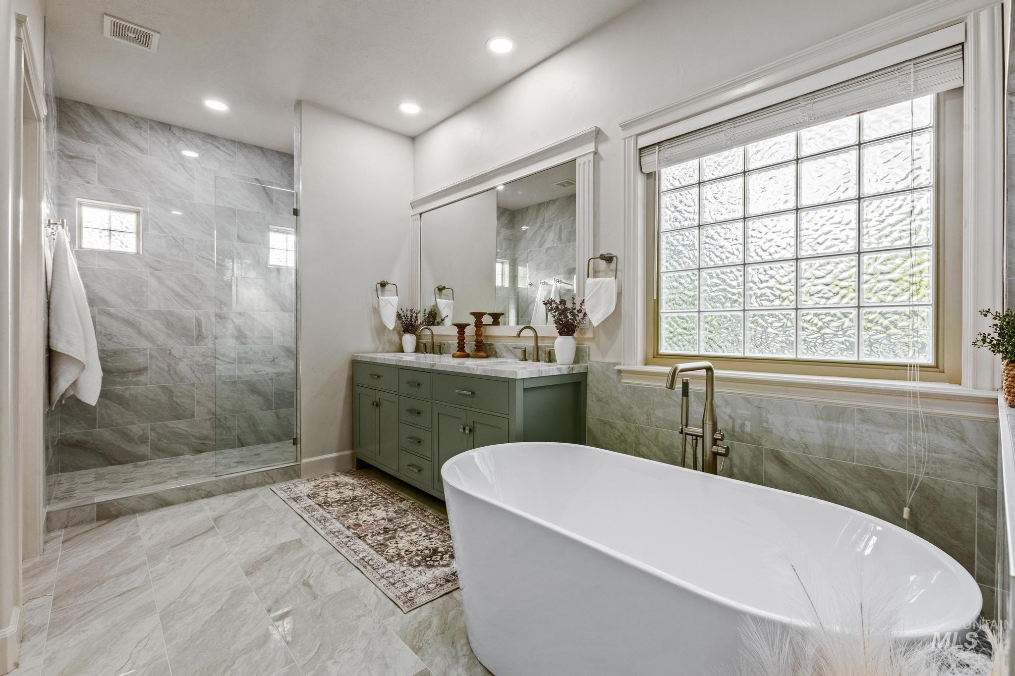 4946 North Villa Ridge Way Boise, ID 83703 - Photo 20 of 49 Spacious bathroom featuring a freestanding soaking tub, a walk-in shower with clear glass enclosure and tile surround, a double vanity with a green cabinet base and stone countertop, and a large window with privacy glass blocks