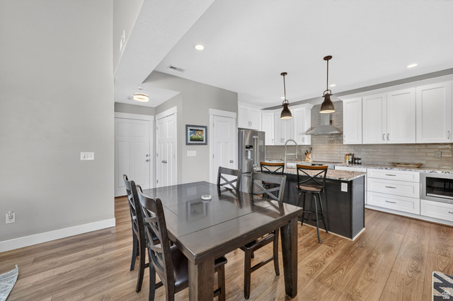 7872 Farm House Lane Midvale, UT 84047 - Photo 8 of 39 Open-concept kitchen and dining area featuring wood-finish flooring, white cabinetry, stainless steel appliances, a center island with bar seating, and recessed lighting