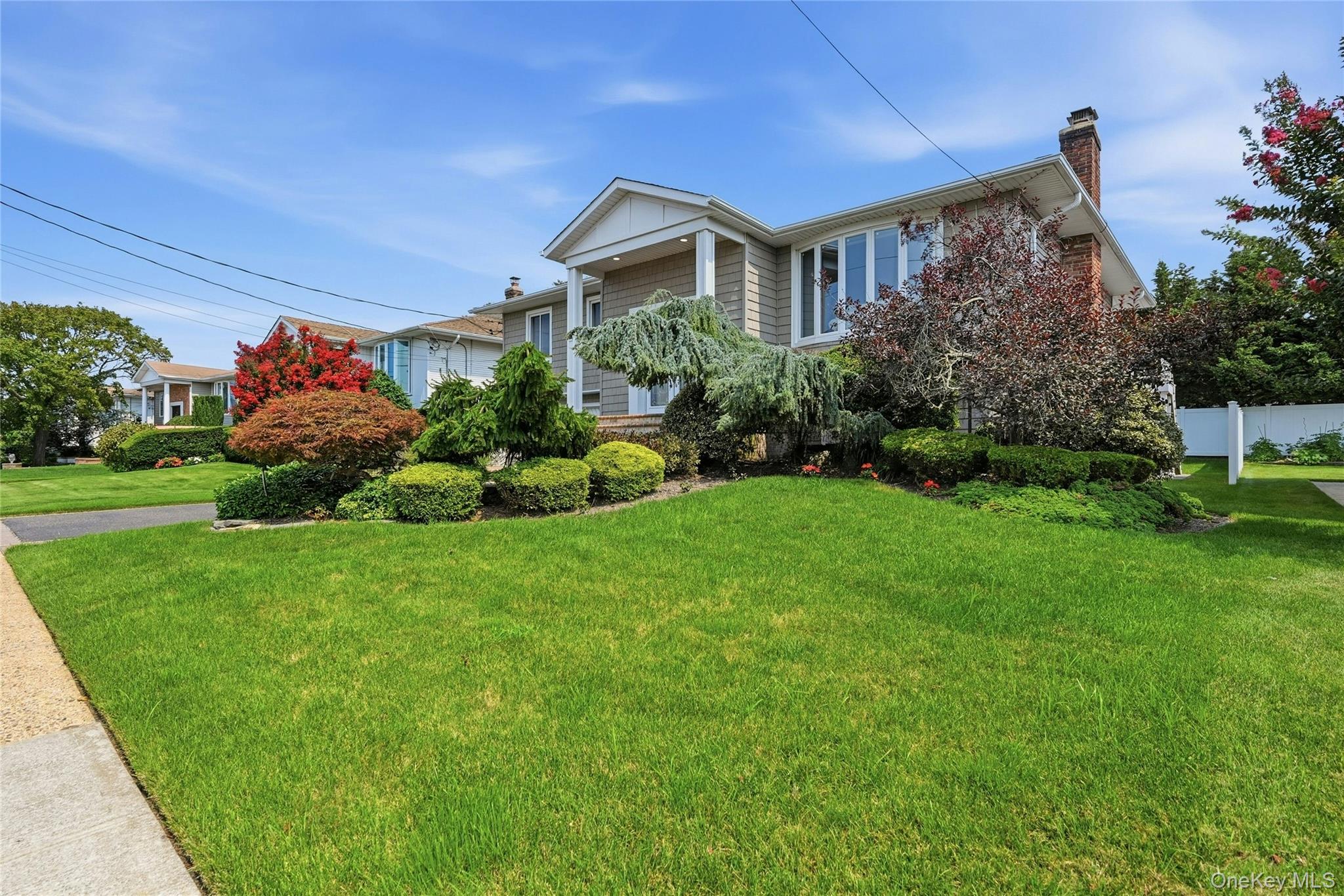 Manicured front lawn with mature landscaping, architectural shingle roof, brick facade, and a white fence