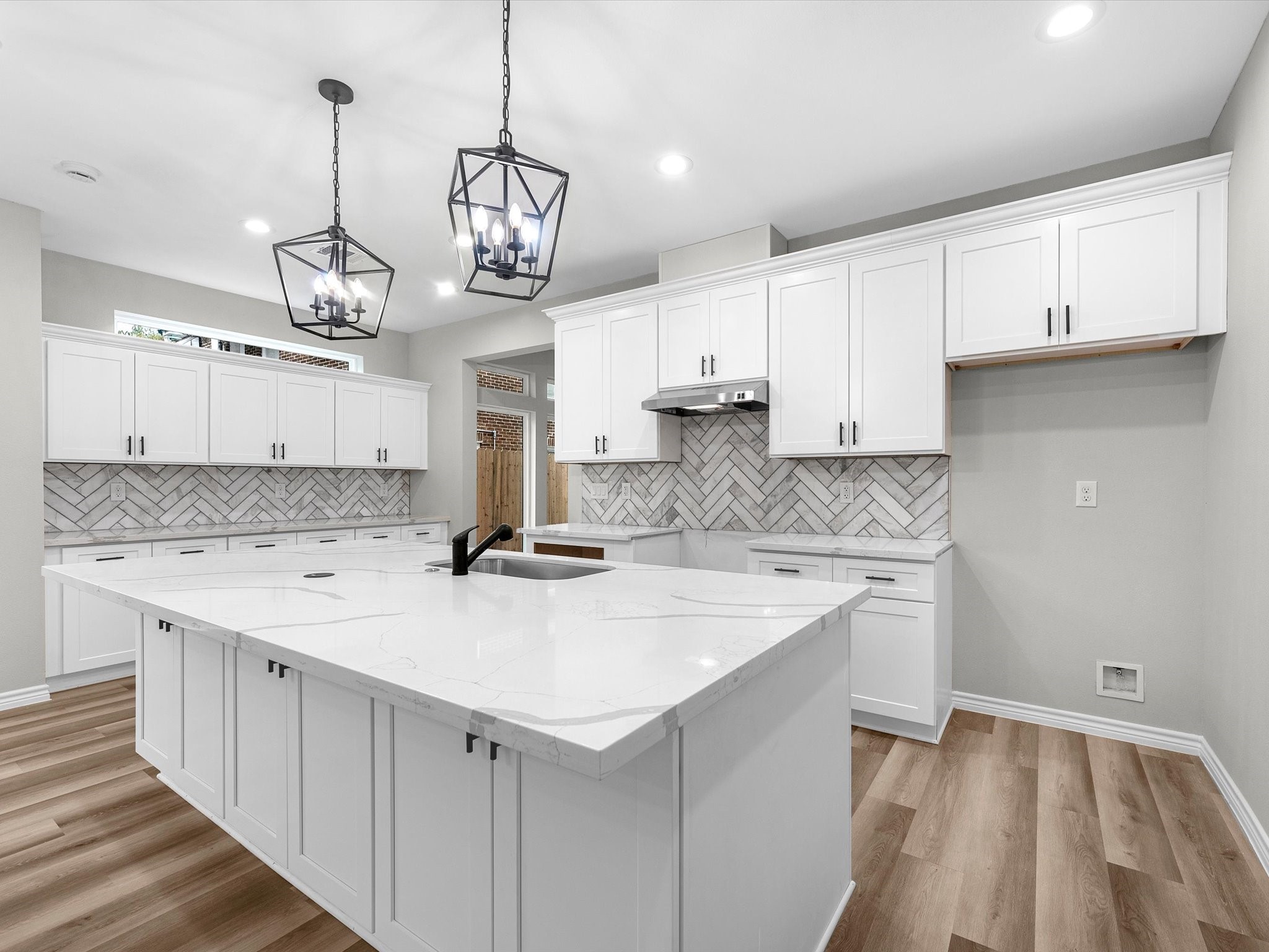 1319 West Bell Street Houston, TX 77019 - Photo 11 of 50 This modern kitchen features a spacious marble-topped island, elegant pendant lighting, and white cabinetry with sleek black handles. The herringbone tile backsplash adds a stylish touch, and the light vinyl flooring enhances the bright, contemporary feel.