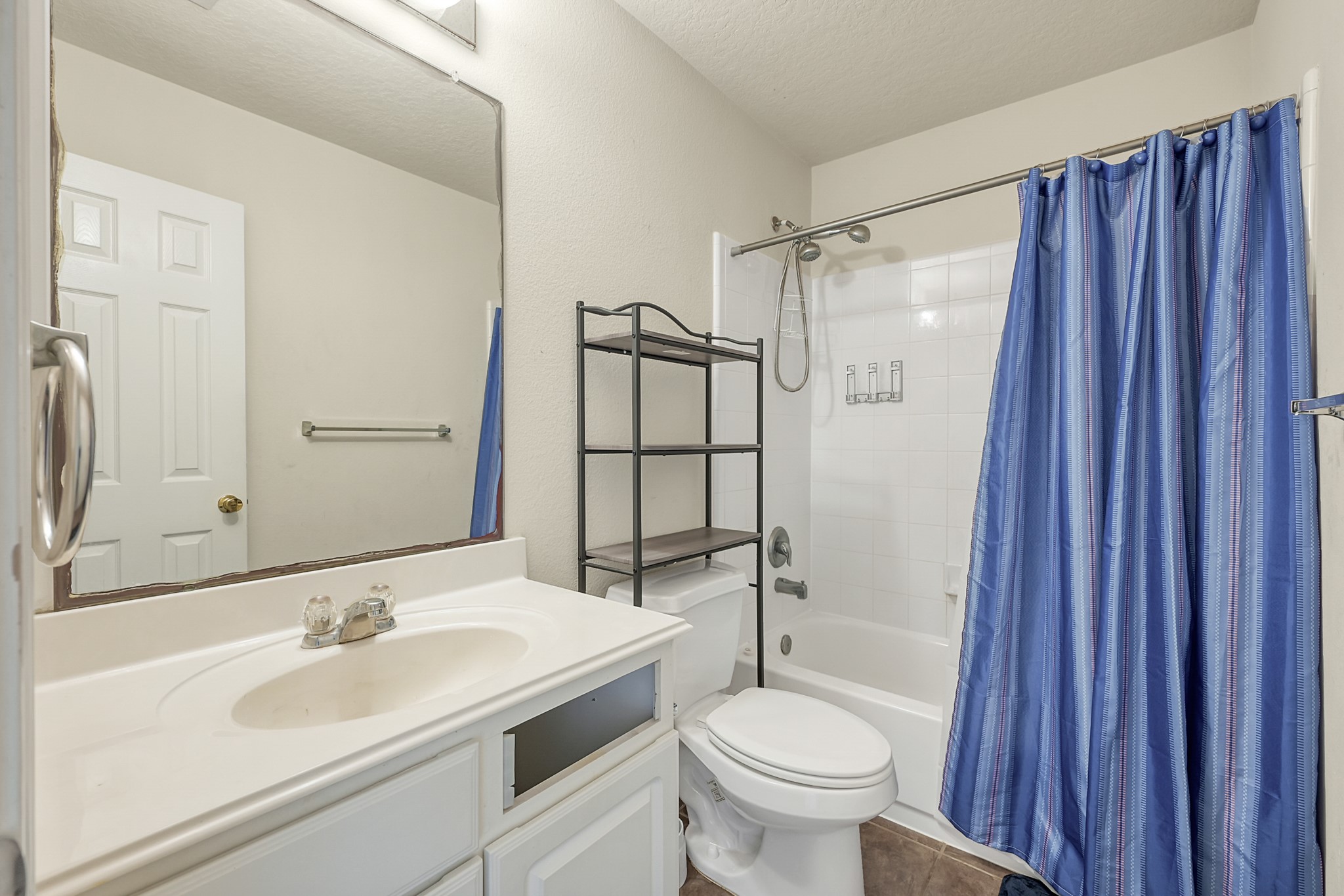22518 Goss Spring Court Spring, TX 77373 - Photo 13 of 19 This bathroom features a clean, white vanity with ample storage, a large mirror, and a shower-tub combo with a bright blue curtain. It includes a metal shelving unit for additional storage and a neutral tile floor.