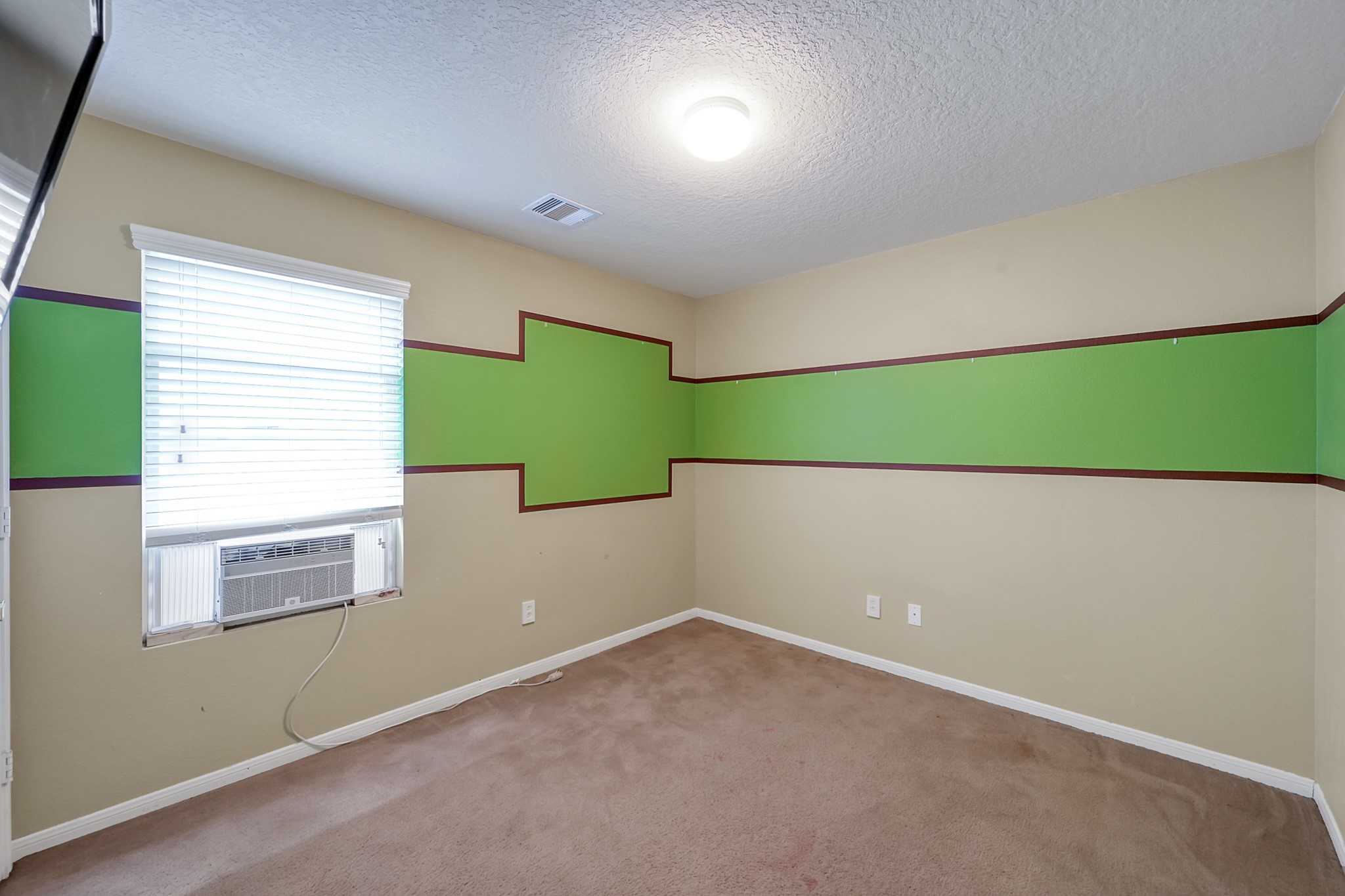 22518 Goss Spring Court Spring, TX 77373 - Photo 14 of 19 This is a small, carpeted room with beige walls featuring a decorative green and brown stripe. It has a window with blinds and a window air conditioning unit. The room is well-lit with a ceiling light.