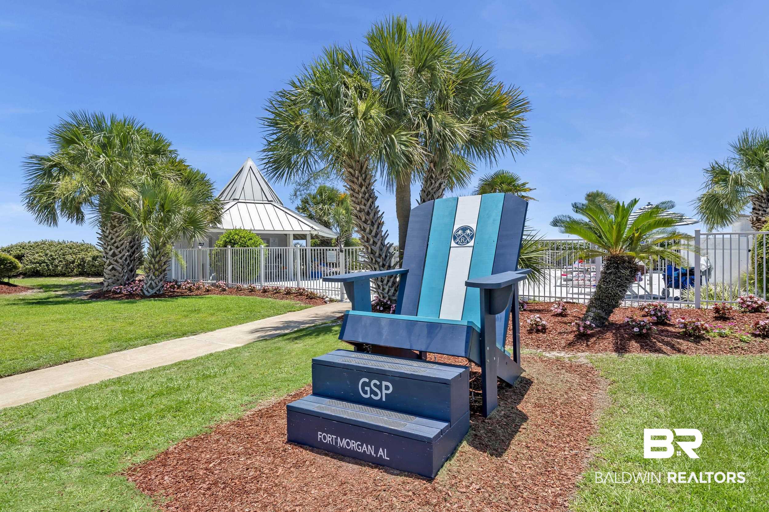 400 Plantation Road, Unit 2118 Gulf Shores, AL 36542 - Photo 26 of 59 Landscaped grounds featuring a large Adirondack-style chair, palm trees, and a concrete pathway leading to a fenced recreational building with a conical roof