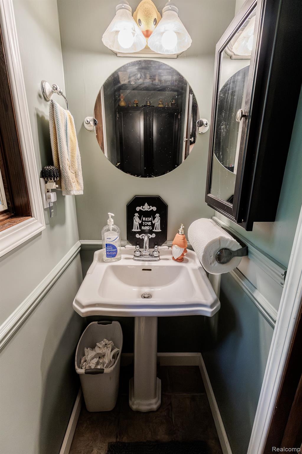 8197 Irish Road Millington, MI 48746 - Photo 31 of 38 Compact powder room featuring a pedestal sink with chrome faucet, an oval wall-mounted mirror, a black medicine cabinet, and a two-light vanity fixture