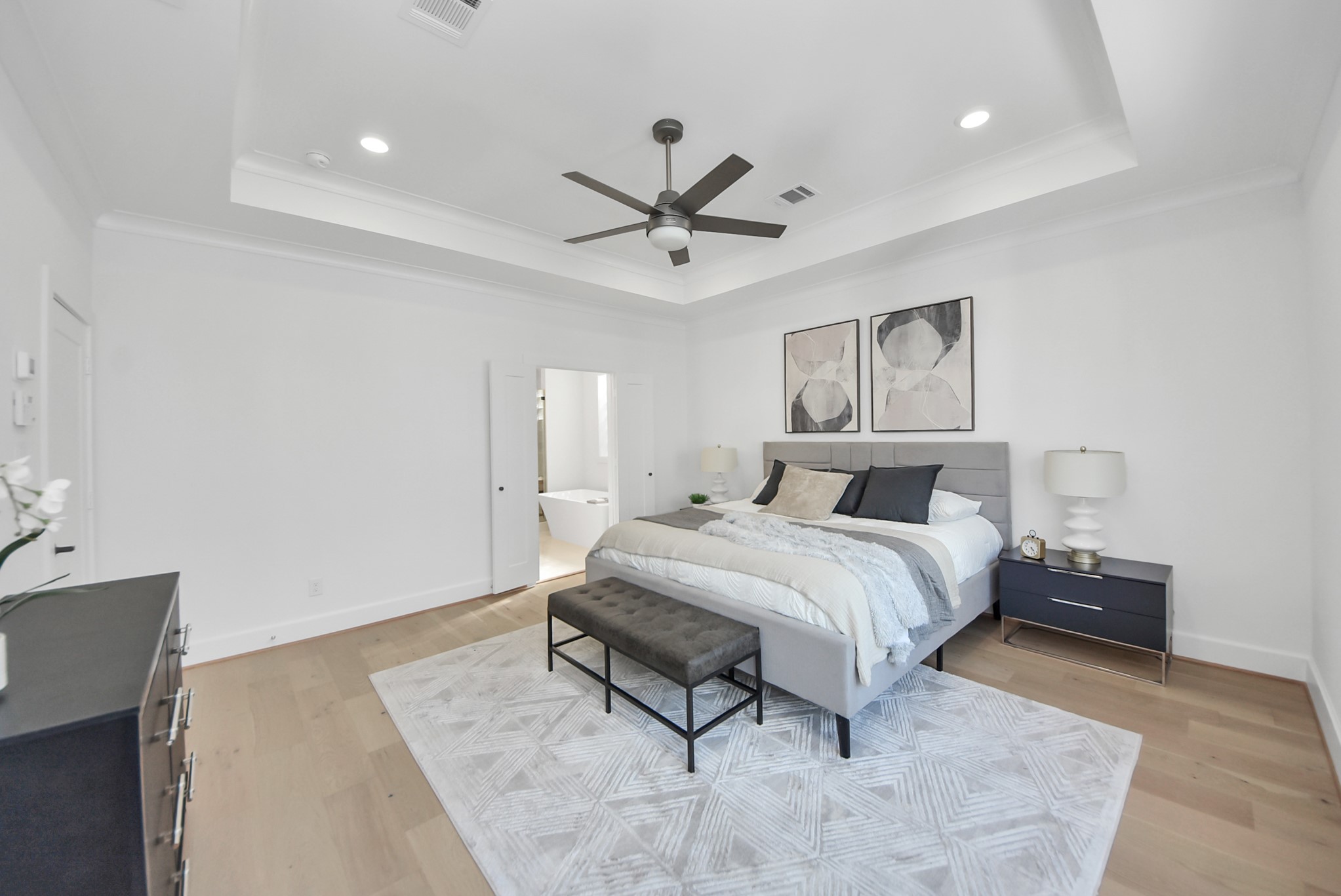 927 Fisher Street, Unit B Houston, TX 77018 - Photo 23 of 47 This spacious, modern bedroom invites relaxation with a neutral color palette, tray ceiling, recessed lighting, and a ceiling fan. A large bed with stylish bedding and matching nightstands anchors the room, while a contemporary rug adds warmth. The room connects seamlessly to an en-suite bathroom, enhancing the suite’s luxurious feel.