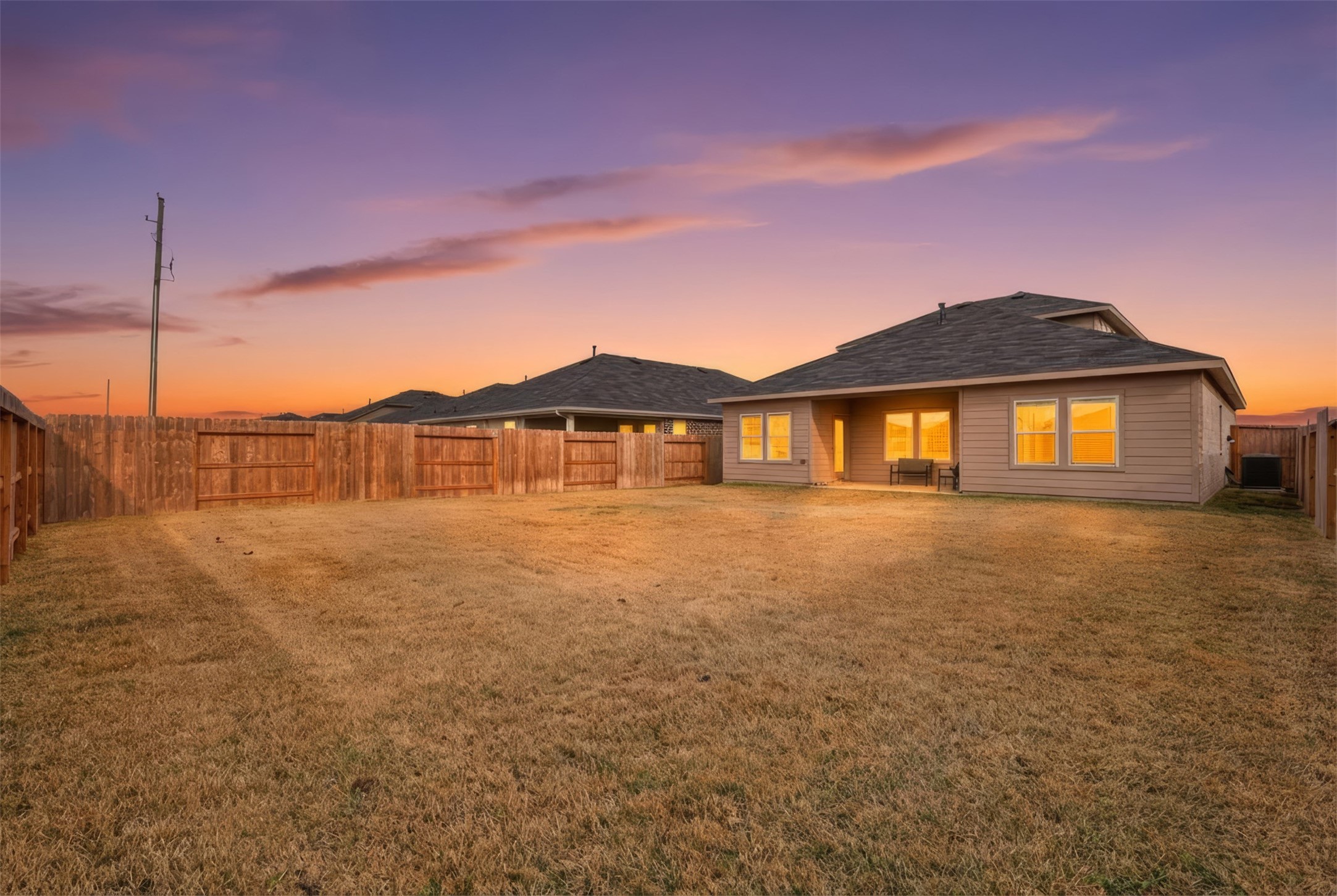 17719 Winfall Drive Crosby, TX 77532 - Photo 40 of 47 Spacious backyard with a well-maintained lawn, enclosed by a wooden fence. The house features a covered patio and large windows, creating a warm, inviting atmosphere during sunset. Ideal for outdoor activities and relaxation.