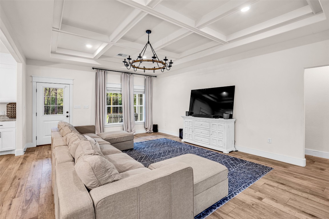 10040 Shepard Hill Road Willis, TX 77318 - Photo 14 of 36 Spacious living room featuring a coffered ceiling, elegant chandelier, and large windows for natural light. It includes a cozy sectional sofa, a large TV on a white console, and a stylish blue rug, all complemented by wood flooring. Perfect for relaxation and entertainment.