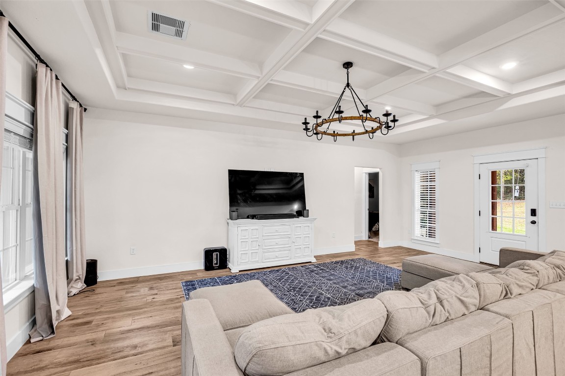 10040 Shepard Hill Road Willis, TX 77318 - Photo 15 of 36 This spacious living room features a coffered ceiling with a stylish chandelier, a large TV on a white console, and a comfortable sectional sofa. The room is bright with natural light from multiple windows and a glass door, complemented by wood flooring and neutral walls.