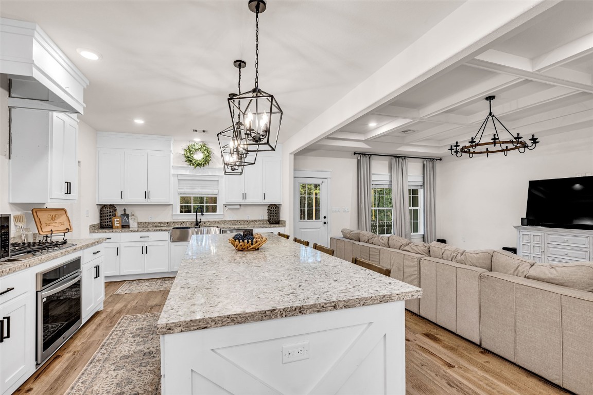 10040 Shepard Hill Road Willis, TX 77318 - Photo 18 of 36 This photo showcases a modern, open-concept kitchen and living room. The kitchen features white cabinetry, granite countertops, and stainless steel appliances, with a large island and pendant lighting. The living area includes a cozy sectional sofa and a wall-mounted TV, complemented by hardwood floors and elegant ceiling fixtures.
