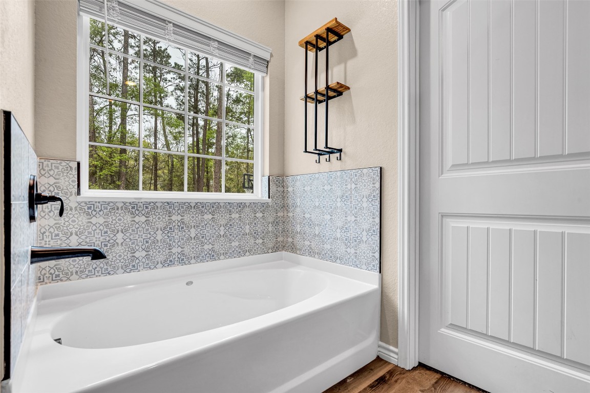 10040 Shepard Hill Road Willis, TX 77318 - Photo 30 of 36 This bathroom features a modern bathtub with intricate tile accents and a large window offering serene forest views. A sleek black faucet complements the design, and there’s a stylish wall-mounted shelf for convenience.