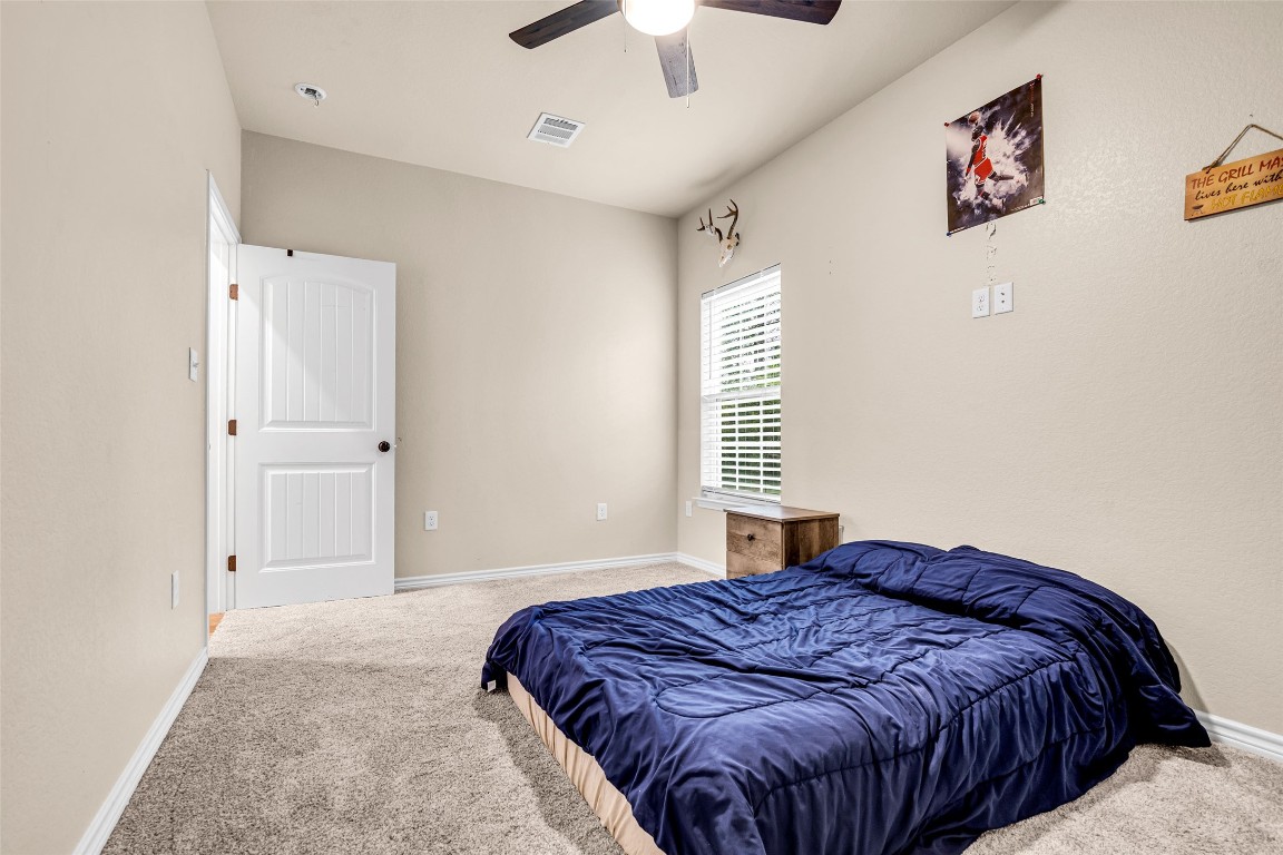 10040 Shepard Hill Road Willis, TX 77318 - Photo 32 of 36 This is a cozy bedroom with beige walls, carpeted flooring, and a ceiling fan. It features a window with blinds letting in natural light, a bed with a dark blue comforter, and some wall decorations, including a small shelf and framed art.