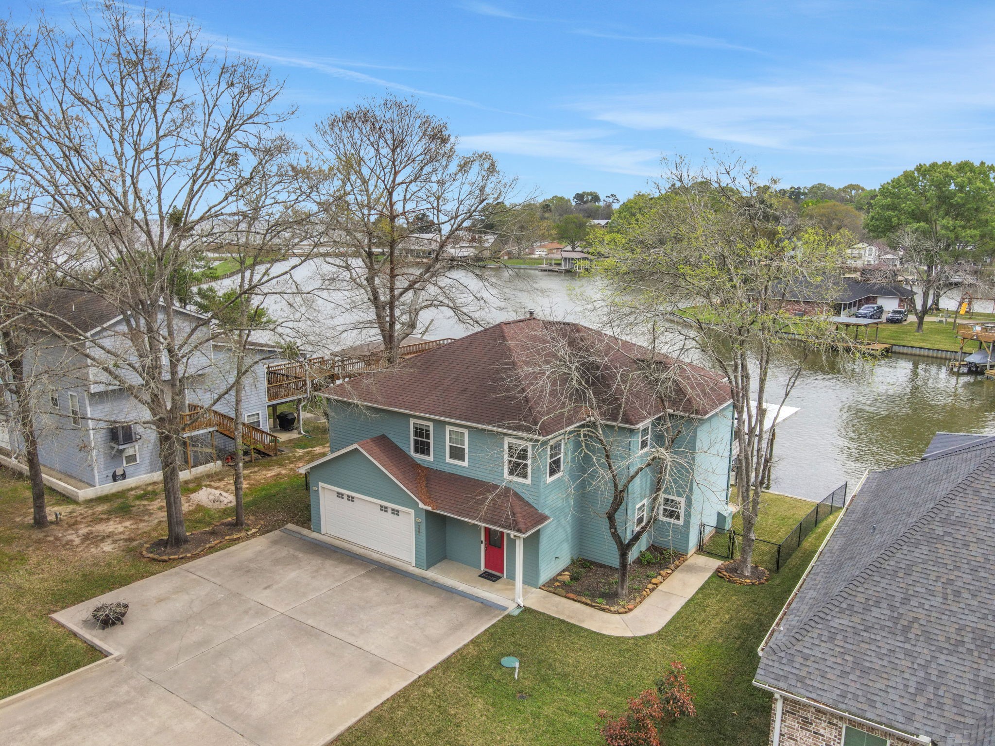 This is a two-story home with a spacious driveway and attached garage, situated on a waterfront lot. The house features a blue exterior with a red front door, surrounded by trees and a neatly maintained lawn. The property offers scenic water views and a serene setting.