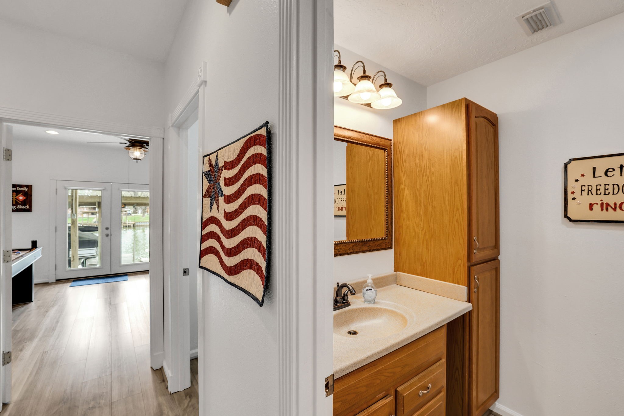 331 Holiday Lane Coldspring, TX 77331 - Photo 12 of 36 This photo shows a bright hallway with light wood flooring leading to a room with large glass doors overlooking water. On the right, there's a small bathroom with a wooden vanity and cabinet.