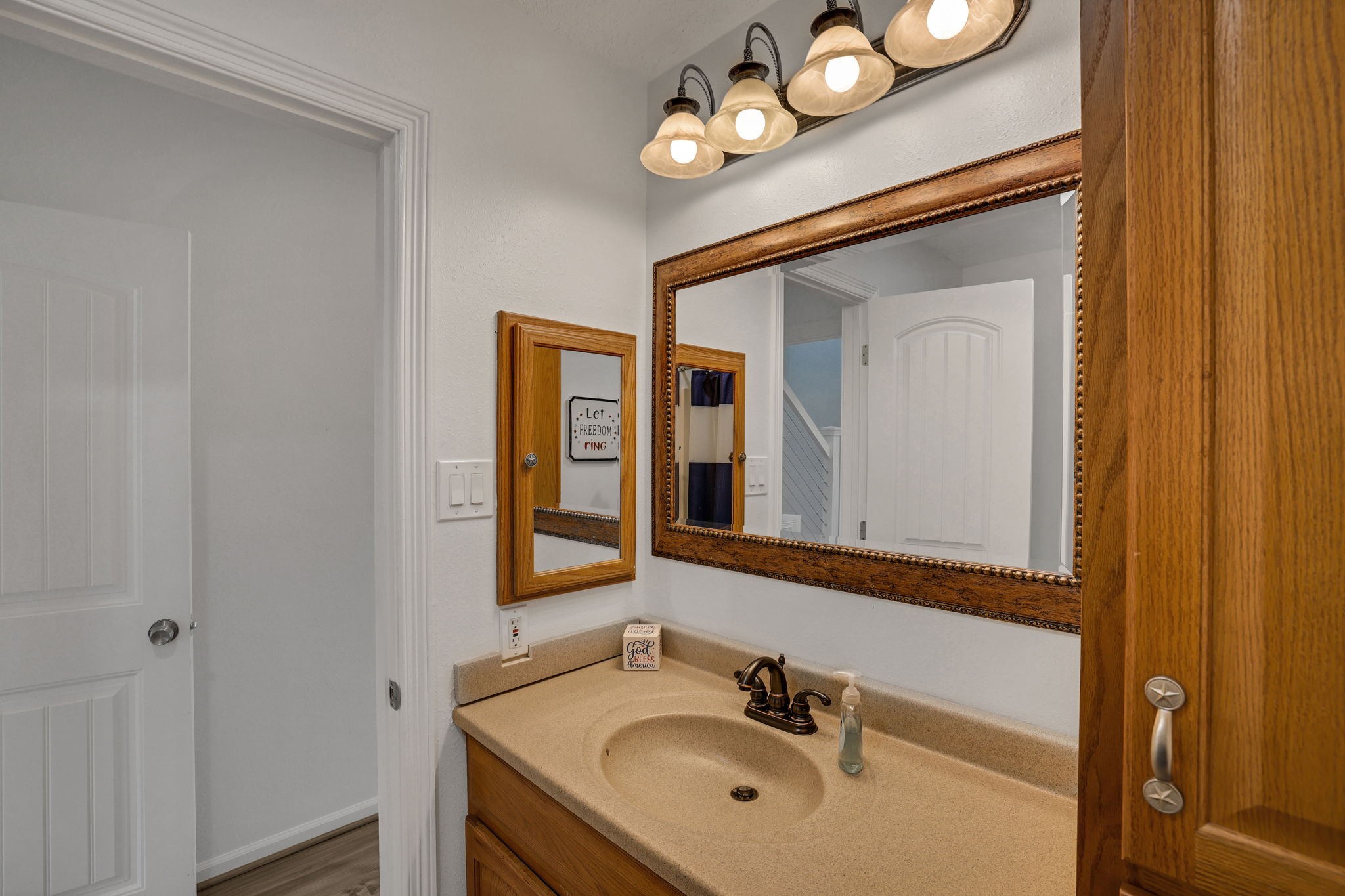 331 Holiday Lane Coldspring, TX 77331 - Photo 25 of 36 This secondary bathroom features a warm, inviting design with wooden cabinetry and a large mirror. It has a beige countertop with an undermount sink and classic bronze fixtures. Above, there are three decorative light fixtures, adding a cozy ambiance.