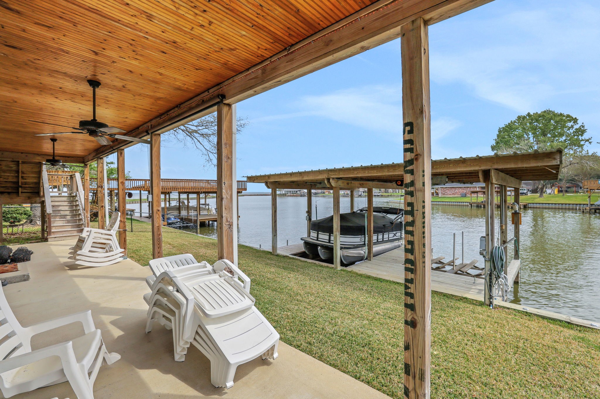 331 Holiday Lane Coldspring, TX 77331 - Photo 26 of 36 This photo showcases a covered patio with a wooden ceiling and ceiling fans, overlooking a serene waterfront. There are stacked lounge chairs and a boat dock, perfect for relaxing and enjoying lake views.