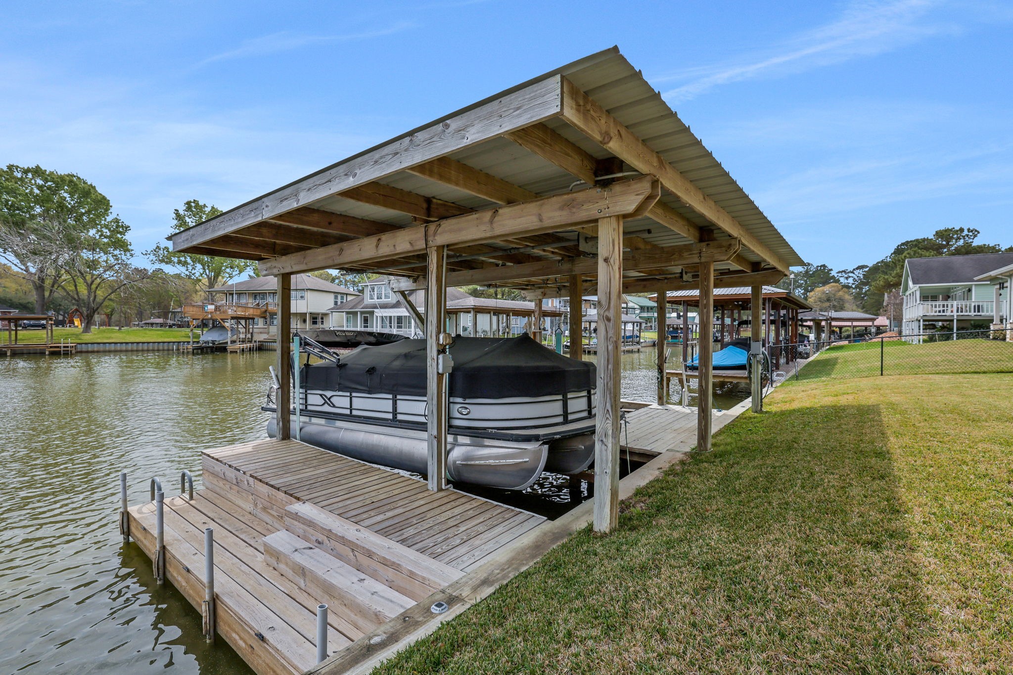 331 Holiday Lane Coldspring, TX 77331 - Photo 29 of 36 This photo shows a waterfront property with a covered boat dock, including a boat lift, set on a peaceful canal. The surrounding area features grassy lawns and neighboring homes, offering a serene and convenient lakeside lifestyle.